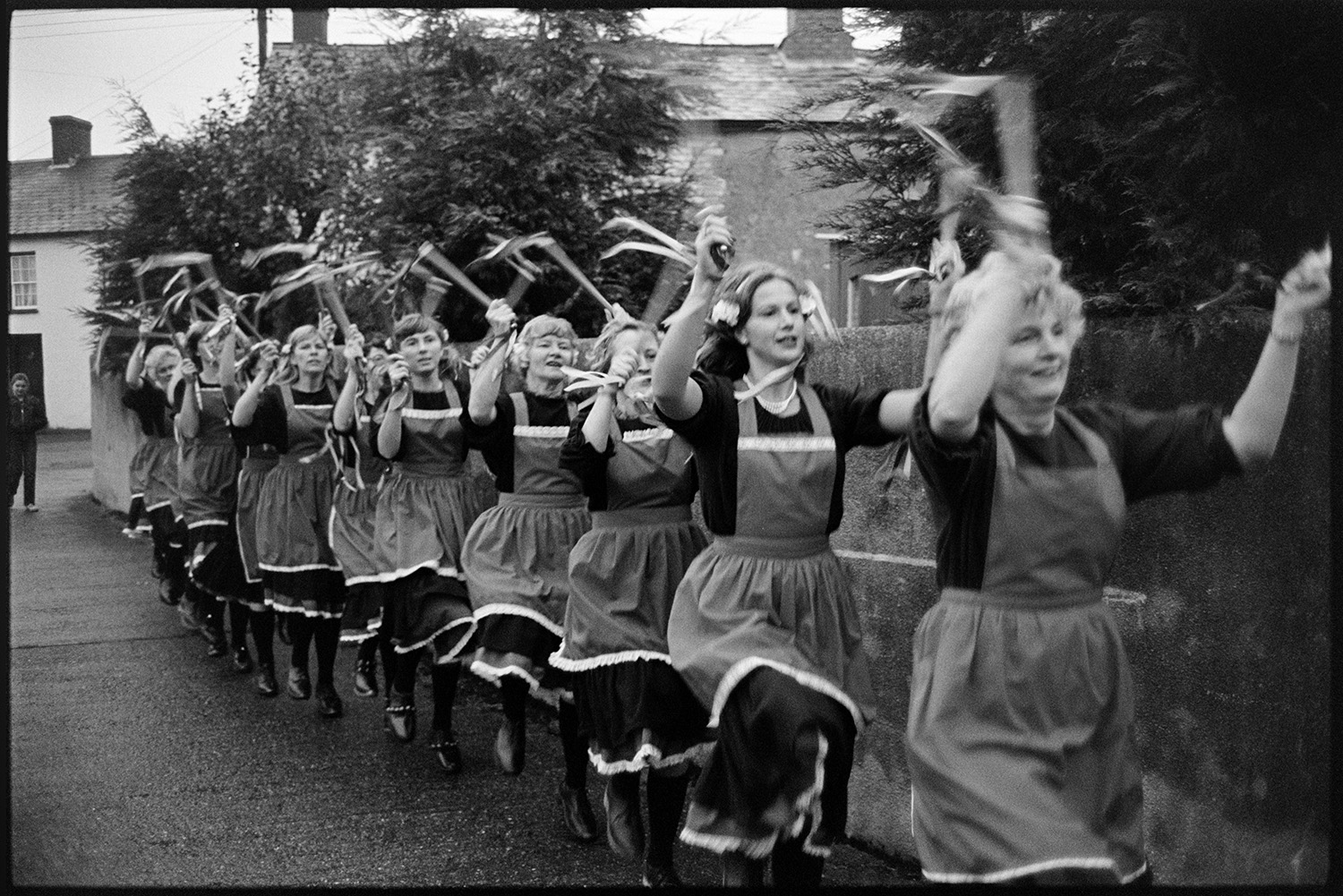 Women's Clog Dancing team at village carnival. 
[A group of women clog dancers performing in a line on a street at Dolton Carnival. They are waving batons with bells.]