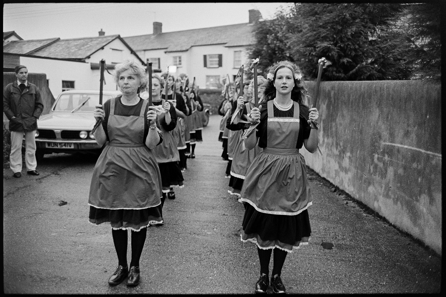 Women's Clog Dancing team at village carnival. 
[A group of women clog dancers in two lines in a street in Dolton, ready to dance in Dolton Carnival. They are holding batons with bells.]