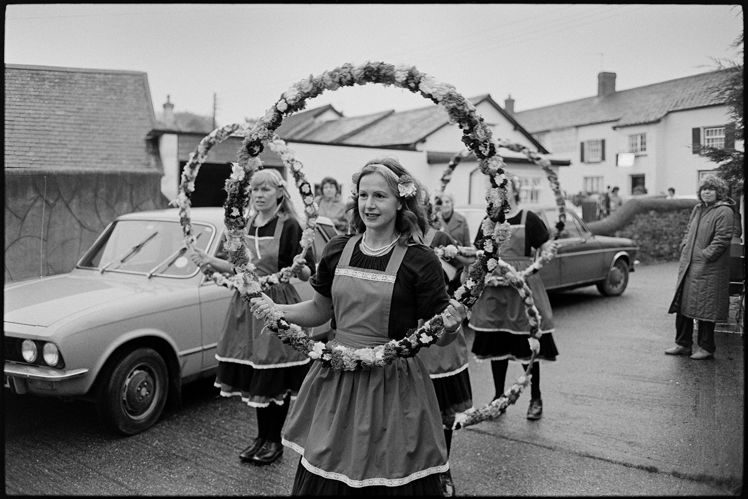 Women's Clog Dancing team at village carnival. 
[A group of women clog dancers performing with floral garlands in a street with parked cars at Dolton Carnival. People are watching them in the background.]