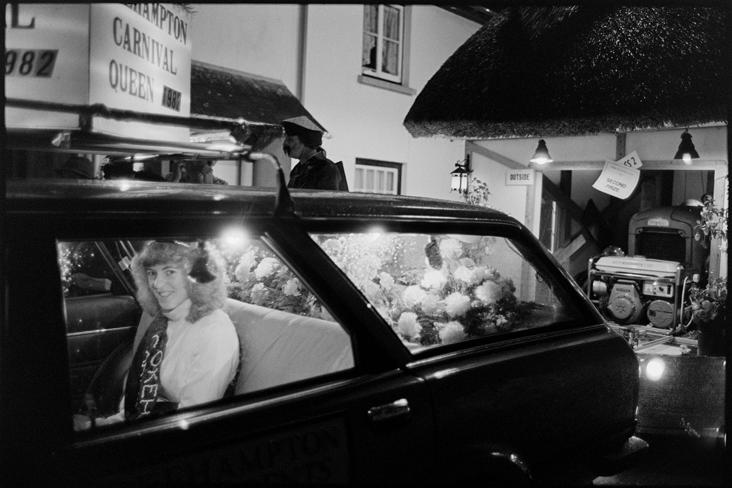 Carnival parade at night, floats flaming torches, Queen in car. 
[The Okehampton carnival Queen in a car with flower arrangement son the parcel shelf, at Dolton Carnival. A thatched garage can be seen in the background.]