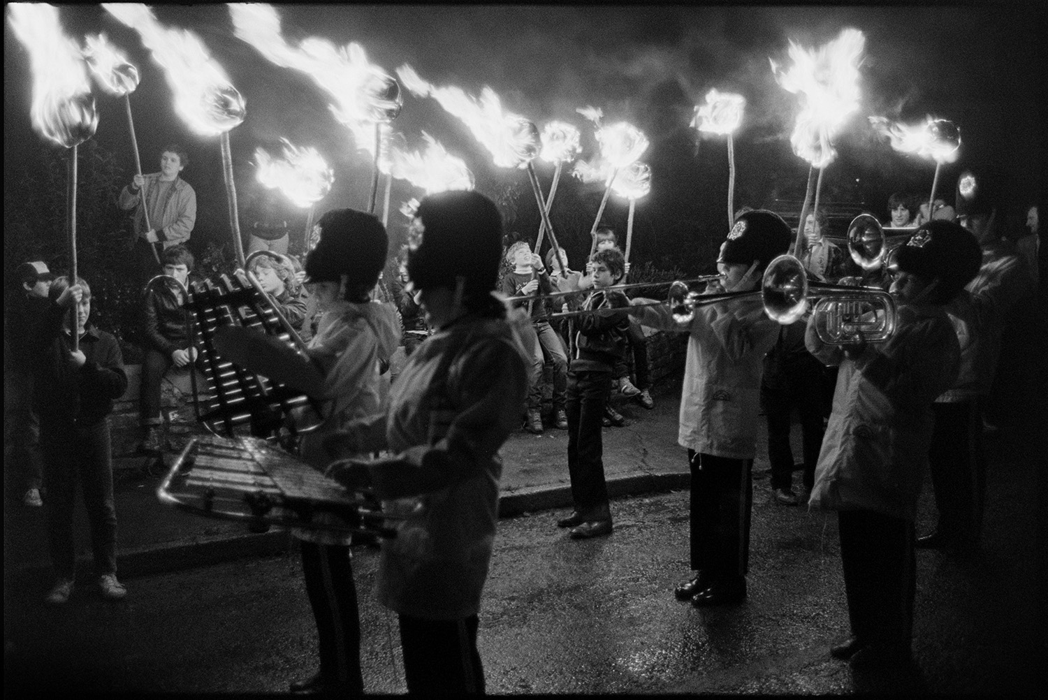 Carnival parade at night, floats flaming torches, Queen in car. 
[A marching band parading along a street at Dolton Carnival. Boys holding flaming torches are lining the street.]
