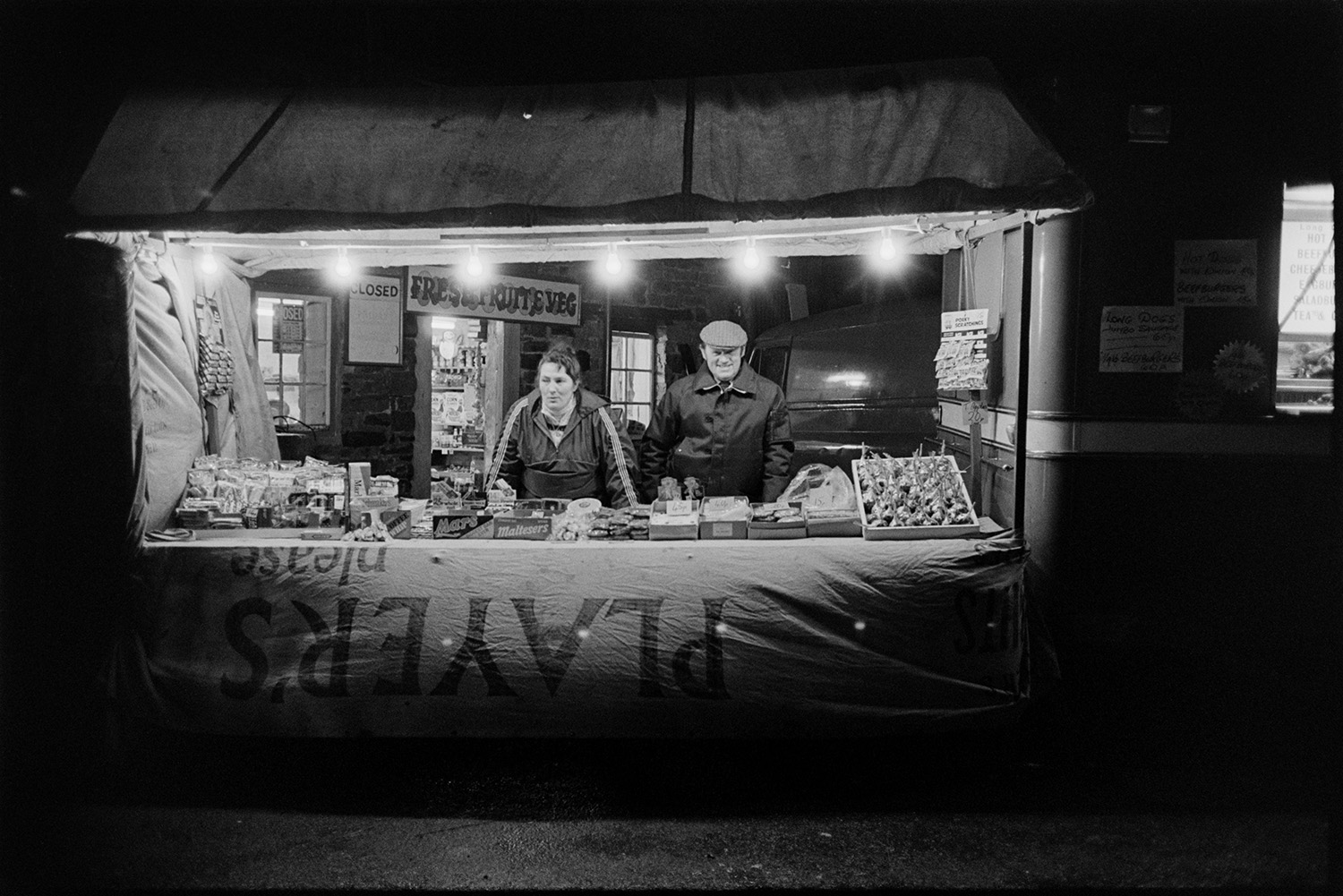 Stall selling hot dogs, sweets and toffee apples at carnival at night. <br />
[Susan Jury and Ken Jury selling toffee apples, sweets and chocolate at a stall at Dolton Carnival at night.]