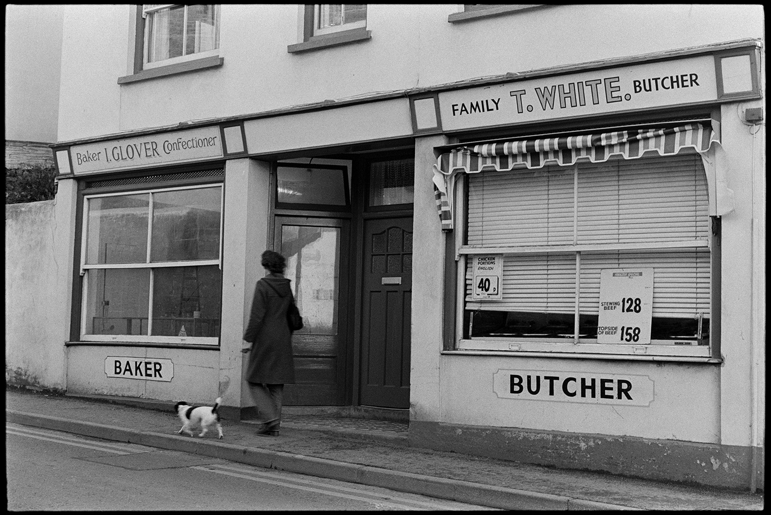 Street scenes with butcher, baker and pub front with sign. 
[A person walking a dog past the shop fronts of T White, butcher and I Glover, Baker, in Bideford.]