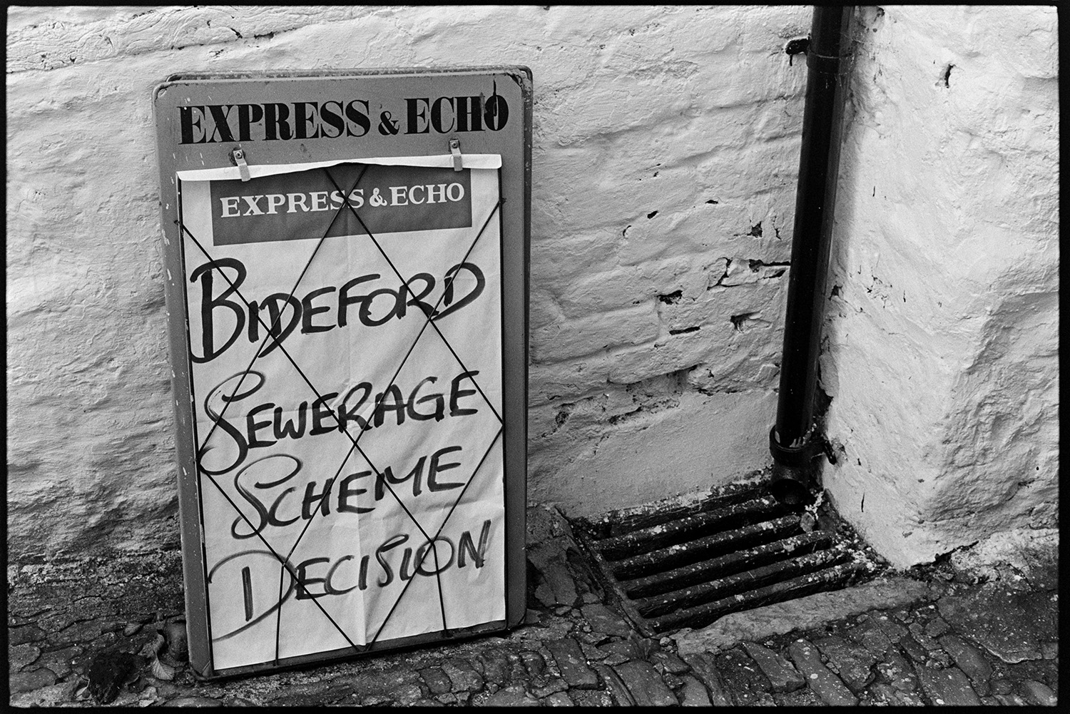 Newspaper hoarding and drain. 
[A newspaper hoarding with the headline 'Bideford Sewerage Scheme Decision' propped against a wall next to a drain with a drainpipe, in Mill Street, Bideford.]