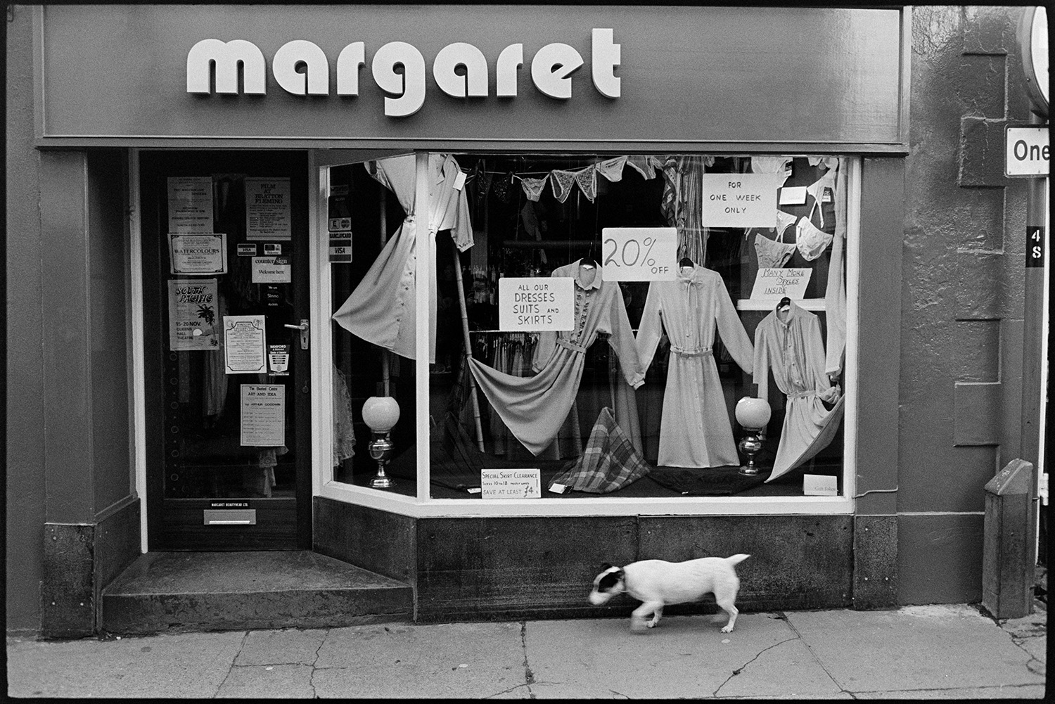 Street scenes with shop fronts, window display of clothes shop. 
[A dog walking past the shop front of 'Margaret' in Mill Street, Bideford. Clothes are displayed in the shop window along with signs advertising 20% off in the shop.]