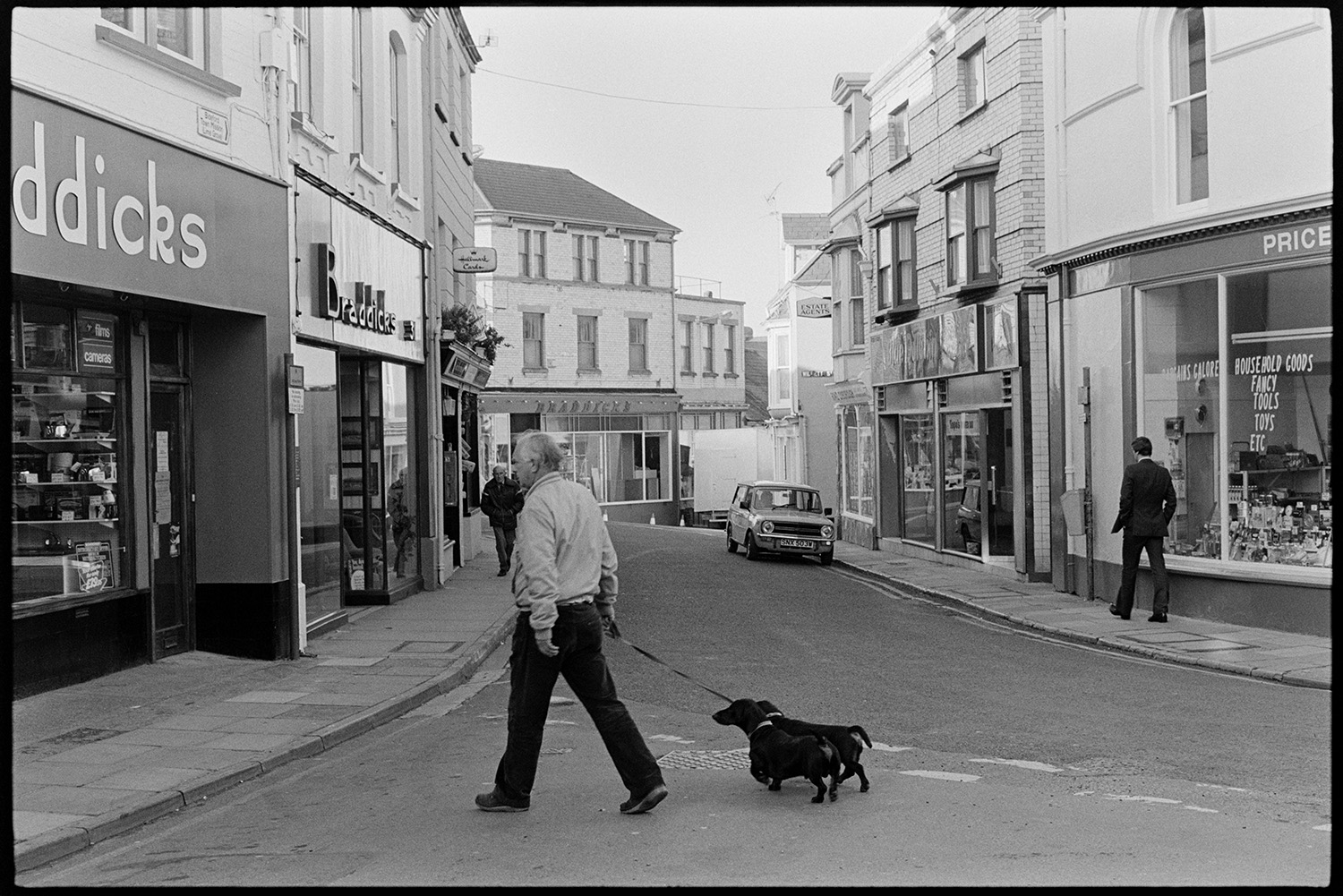 Street scenes with shop fronts, window display of clothes shop. 
[A man crossing Mill Street, Bideford with two dogs. Various shop fronts are visible, including Braddicks.]