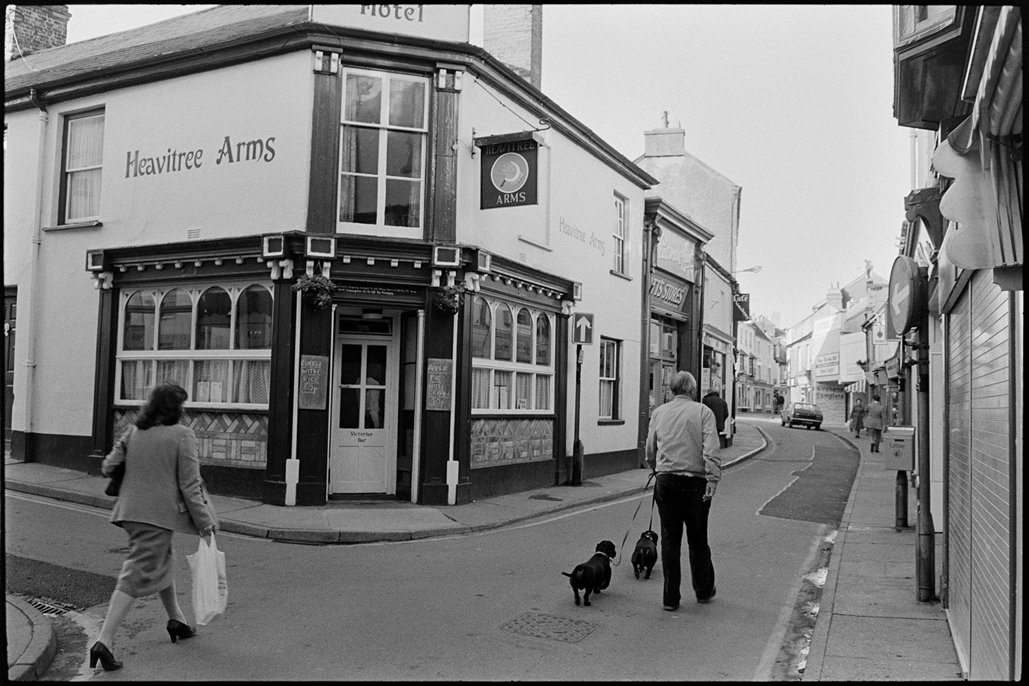 Street scenes with shop fronts, window display of clothes shop. 
[People walking past the entrance to the Heavitree Arms pub in Mill Street, Bideford. One man is walking two dogs. Shop fronts can be seen further along the street.]