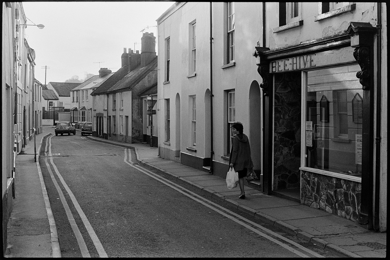 Street scenes with butcher, baker and pub front with sign. 
[A woman walking past the shop front of 'Bee-Hive' in Bideford. Houses and cars can be seen further down the street.]