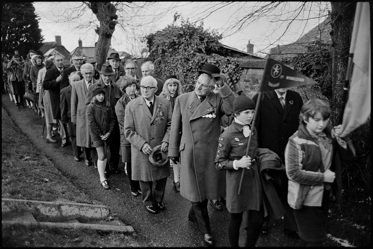Armistice day parade, women and men, old soldiers in church and parade past memorial, wreaths. 
[Men, women and children walking along the path of Winkleigh Church on Remembrance Day. Some of the children are holding standards or flags. Some of the men are wearing medals on their coats.]