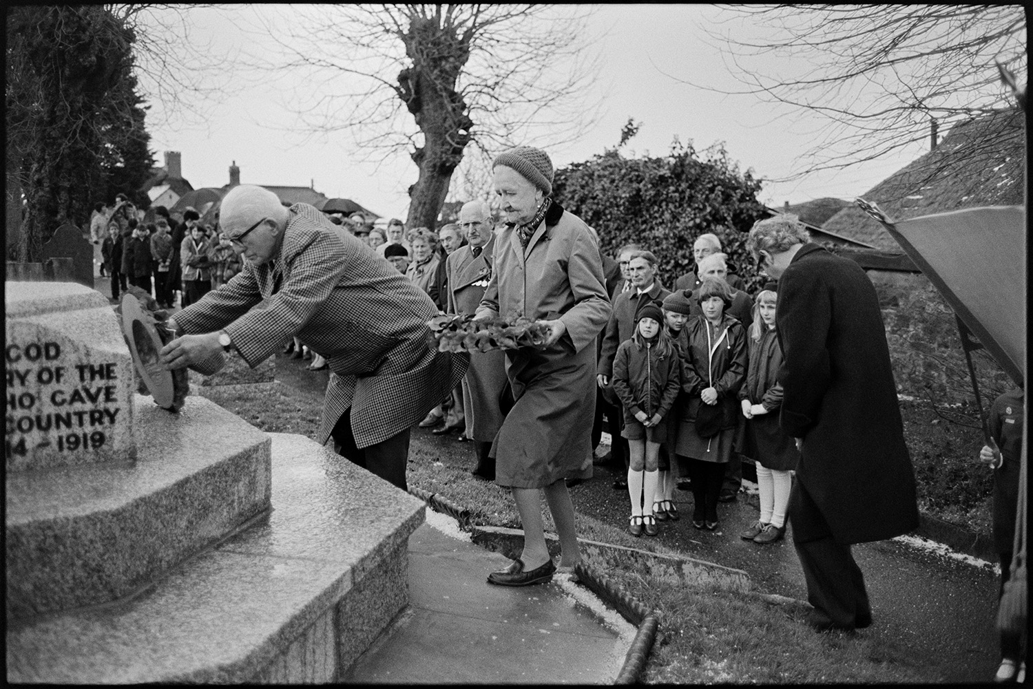 Armistice day parade, women and men, old soldiers in church and parade past memorial, wreaths. 
[A man and a woman laying poppy wreaths at the war memorial in Winkleigh on Remembrance Day. Men, women and children are watching.]