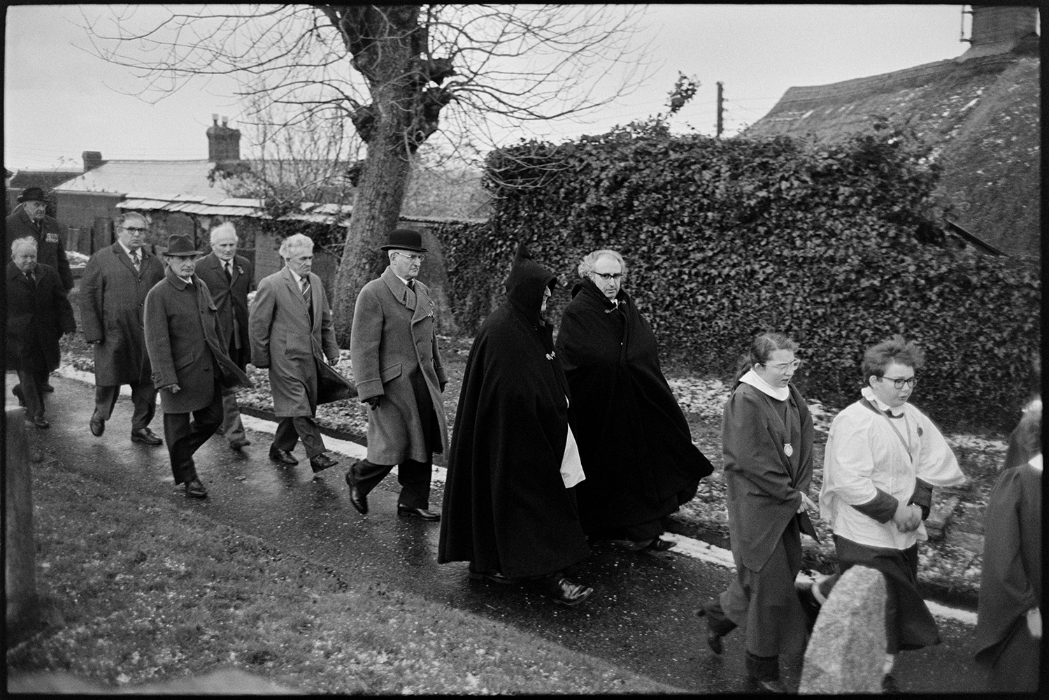 Armistice day parade, women and men, old soldiers in church and parade past memorial, wreaths. 
[Choristers and men walking along the path at Winkleigh church past the war memorial on Remembrance Day. Two men, possibly clergy, are wearing capes. The ground is covered with hail stones.]