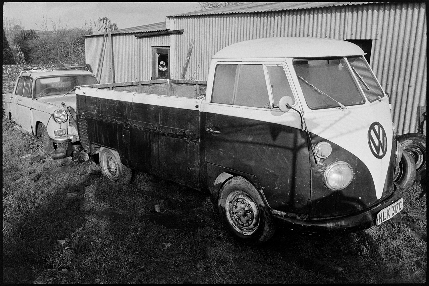 Van, pick up in mucky yard. 
[A Volkswagen pick up van and a car parked by a corrugated iron barn in a muddy yard.]