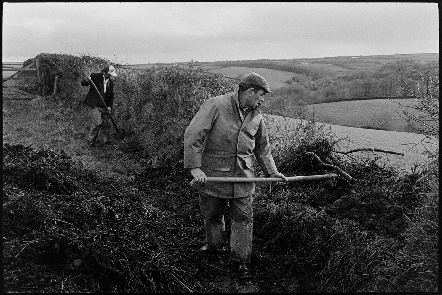 Farmers repairing hedge bank, clatting. 
[Two men clatting  a hedge (building up the hedge with turf) in a field at Narracott, Hollocombe. A landscape of fields and trees can be seen in the background.]