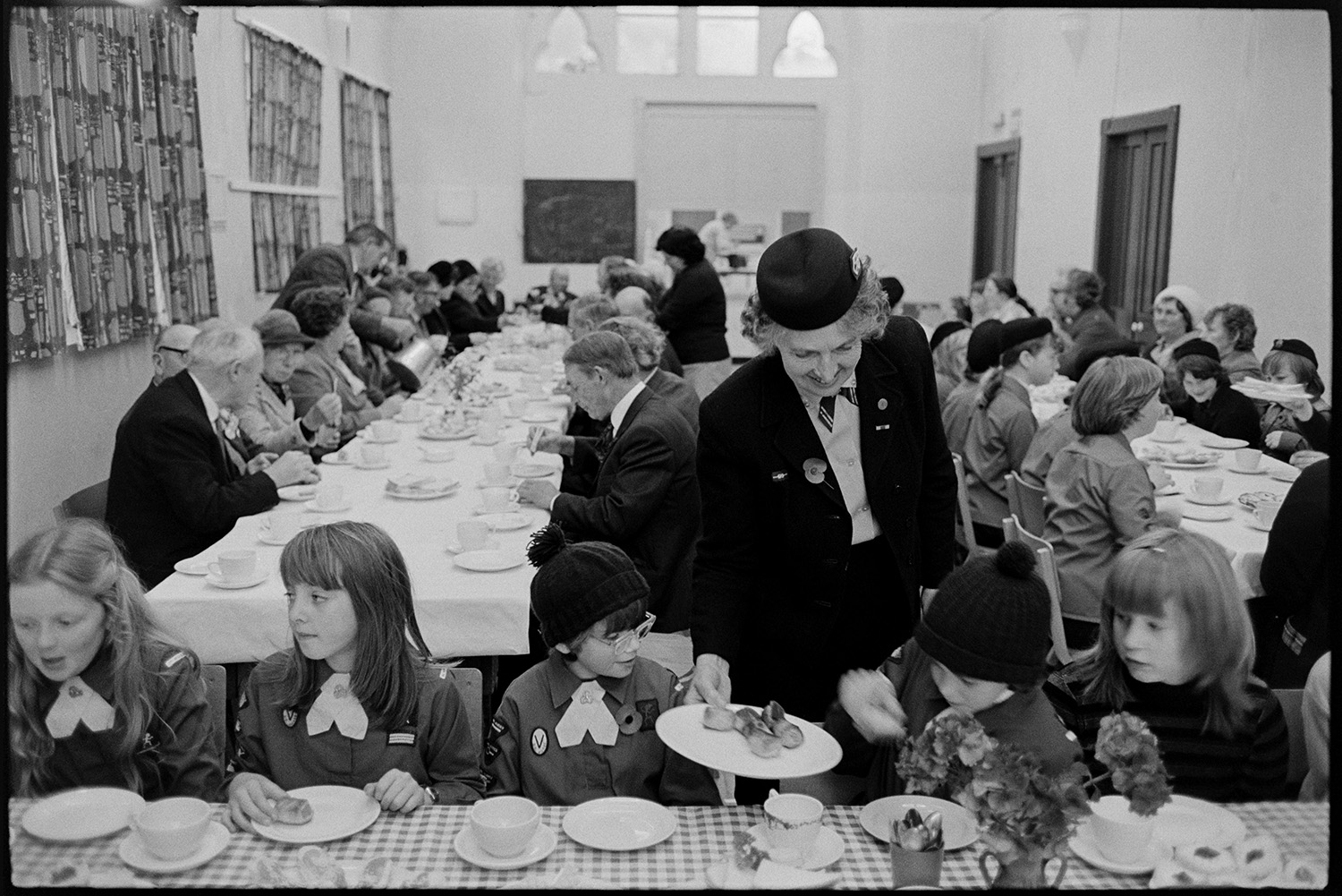 Tea in village hall after Armistice Day parade. 
[Men, women and children having tea in Winkleigh Village Hall after a parade for Remembrance Day. A woman is serving sausage rolls to girls sat at a table in the foreground. The girls are wearing Brownie uniforms.]