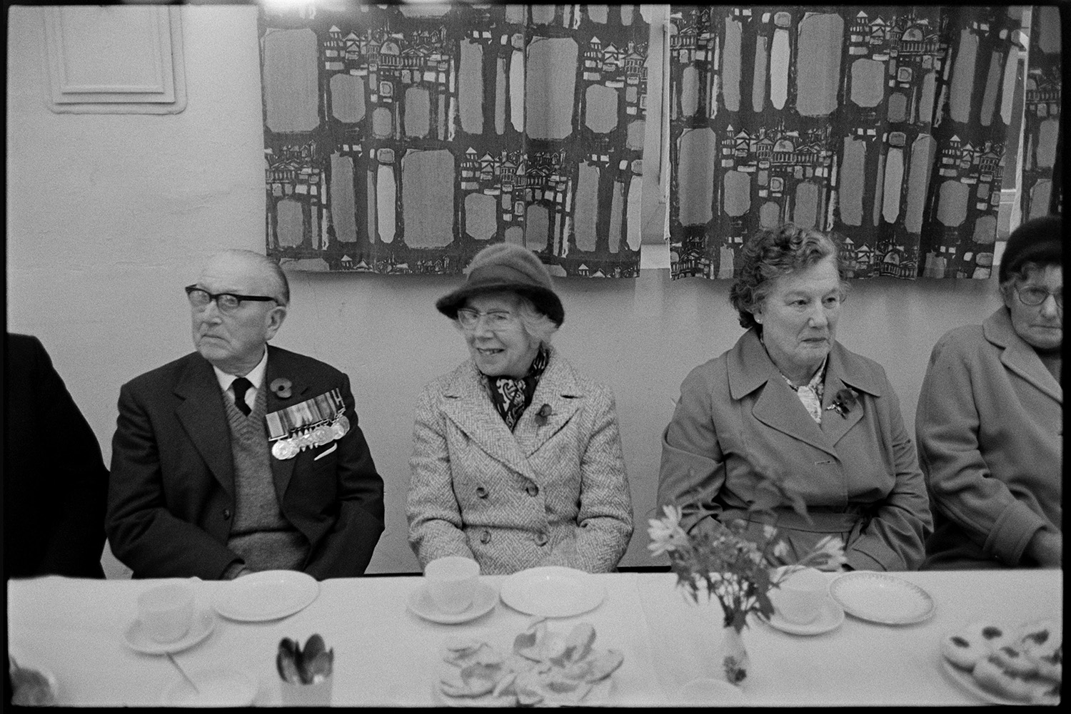 Tea in village hall after Armistice Day parade. 
[A man and three women sat at a table in Winkleigh Village Hall for a tea after the Remembrance Day parade. The table is laid with plates of cakes and the man is wearing military medals on his jacket.]