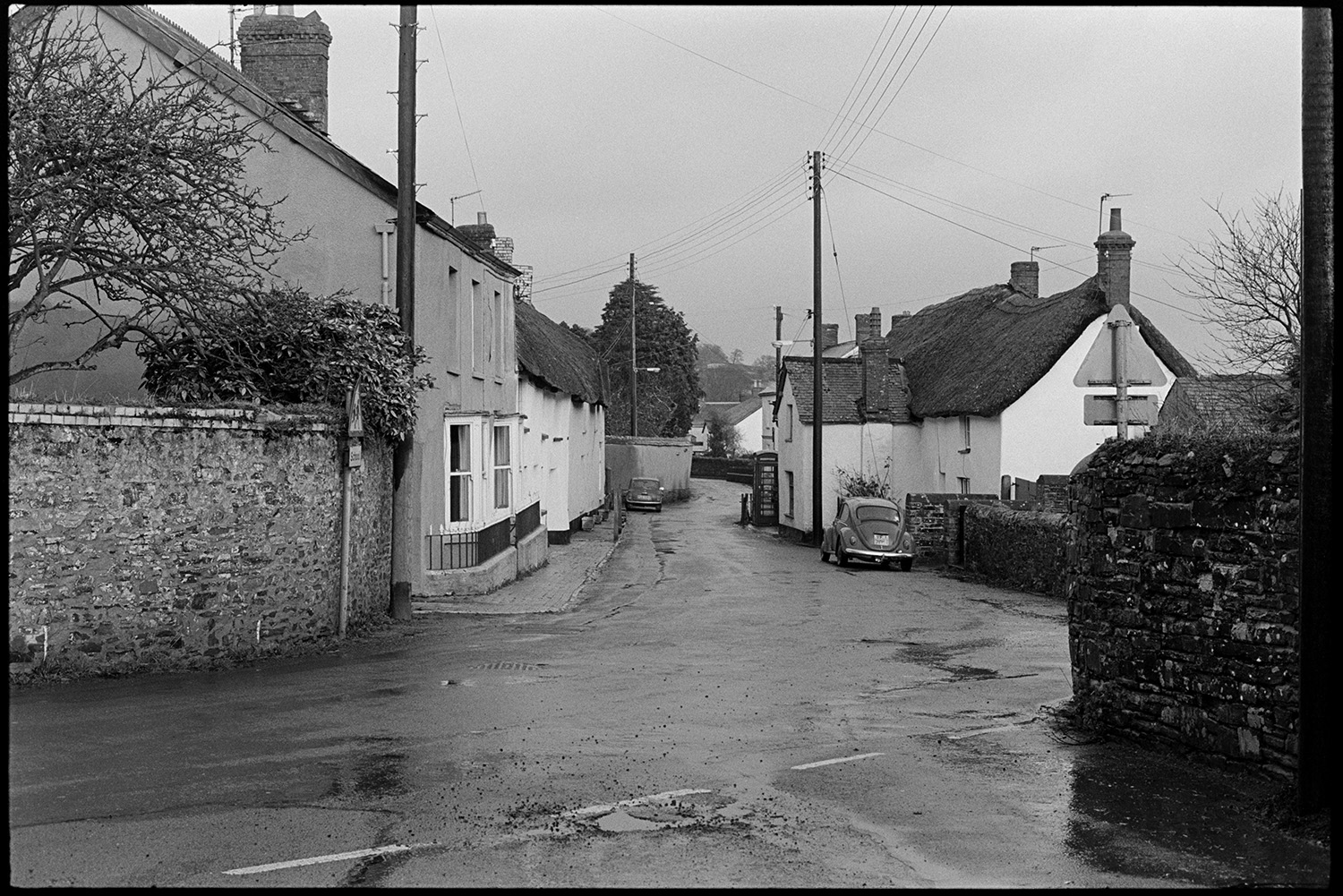 Street scenes, lens test. 
[A view of Fore Street, Dolton with thatched cottages, parked cars and a telephone box. The image was taken during a lens test by James Ravilious.]