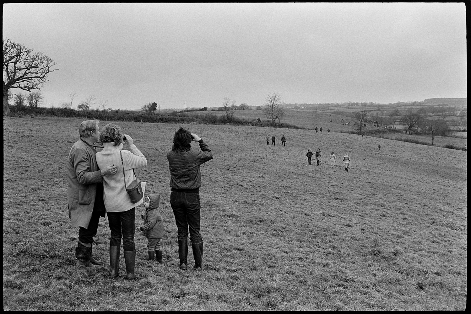 Hunt Meet, horses and followers setting off. 
[Hunt followers walking across a field at Highampton. Two women are watching the hunt in the distance through binoculars in the foreground. A man and child are with them.]