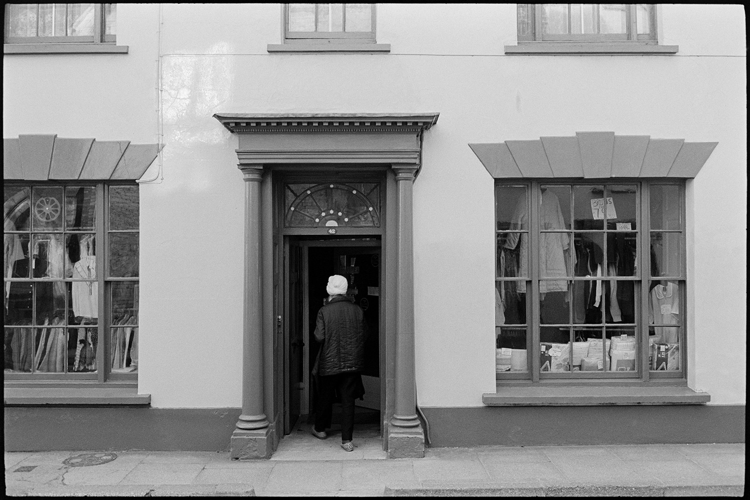 Front of clothes shop. 
[A person walking into Singh's clothes shop in South Street, Torrington. Clothes are displayed in the shop front windows.]