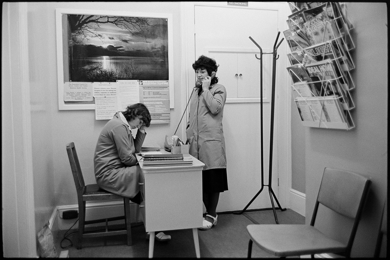 Women working in dentists reception, telephoning, sitting at desk. 
[Two women working in a Mr Rollinson's dentist reception, in Halsdon Terrace, Torrington. One woman is sat at a desk and the other is using the telephone by a coat stand. A picture of a lake is hung up behind the desk and magazines are displayed in a rack attached to the wall in the foreground.]