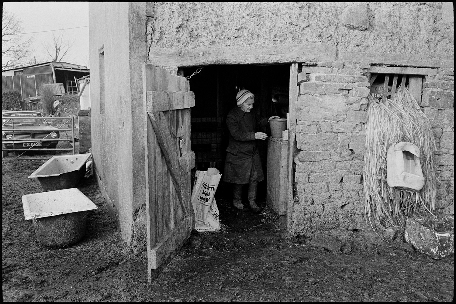 Farmers in milking parlour, churn. 
[Evelyn Folland in the doorway to a stone and cob barn at Higher Upcott, Dowland. Twine is hanging from a window of the barn, and two bath tubs are in the muddy farmyard outside the barn. A car is parked by a gate in the background.]