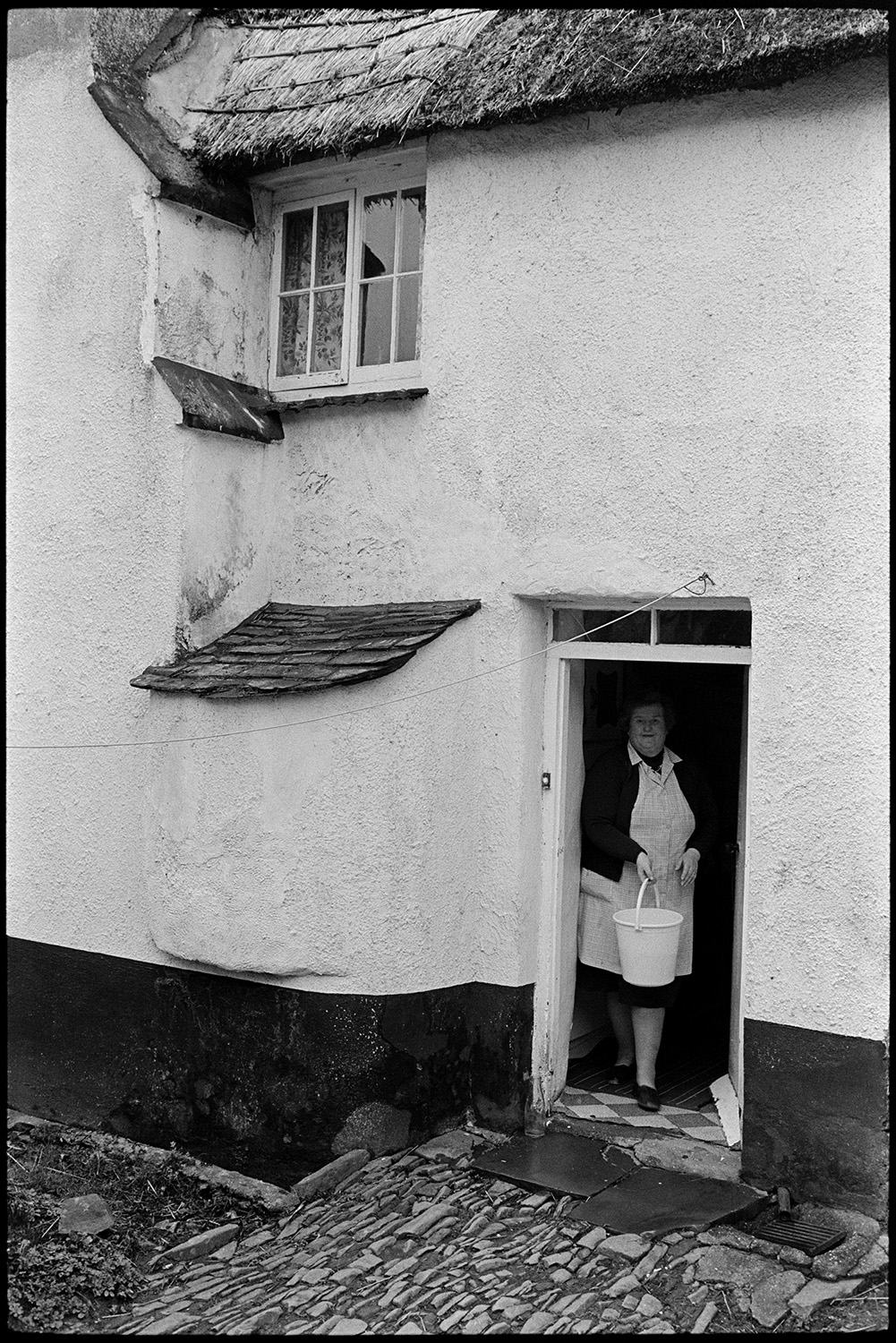 Woman pouring water down drain at back of cob and thatch cottage, see later pic ruined.
[A woman holding a bucket stepping out of the back door of a house in Beaford on to a cobbled path outside to empty the bucket. The cottage has a thatched roof and a tiled bread oven is next to the door.]