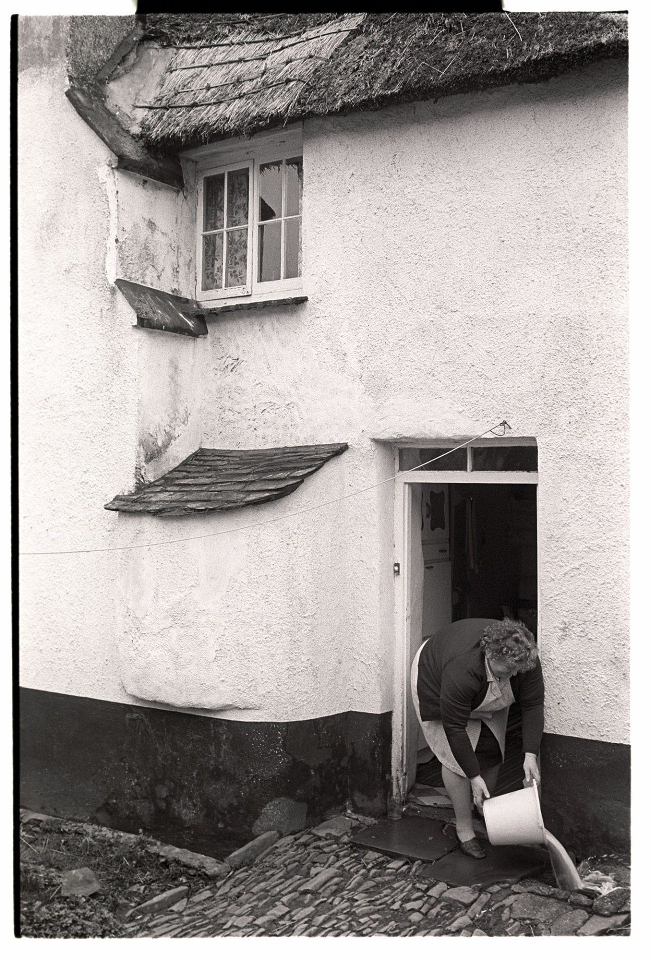 Woman pouring water down drain at back of cob and thatch cottage, see later pic 2080-12. 
[A woman empting a bucket of water down a drain by cobbles at the back of a cob and thatched cottage in Beaford. See also reference RAV/01/2080/12,]
