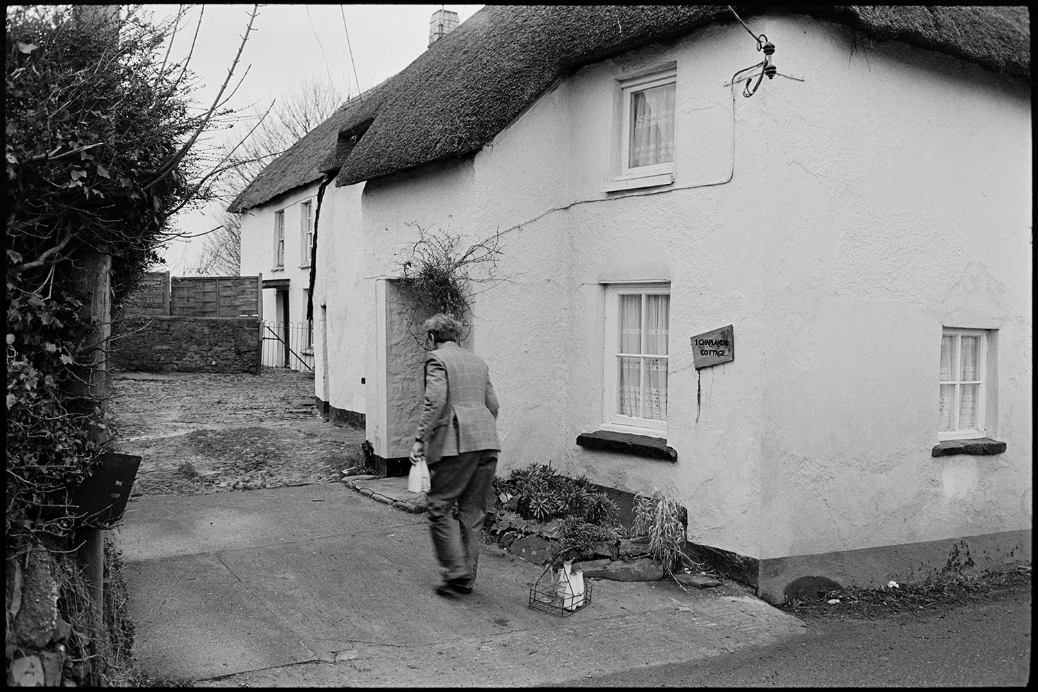 Farmer, milkman doing last milk round, showing new man the ropes, milk bottles.
[Ivor Bourne of Jeffrys farm delivering milk on his last milk round to Chaplands Cottage, Beaford. The cottage has a thatched roof.]