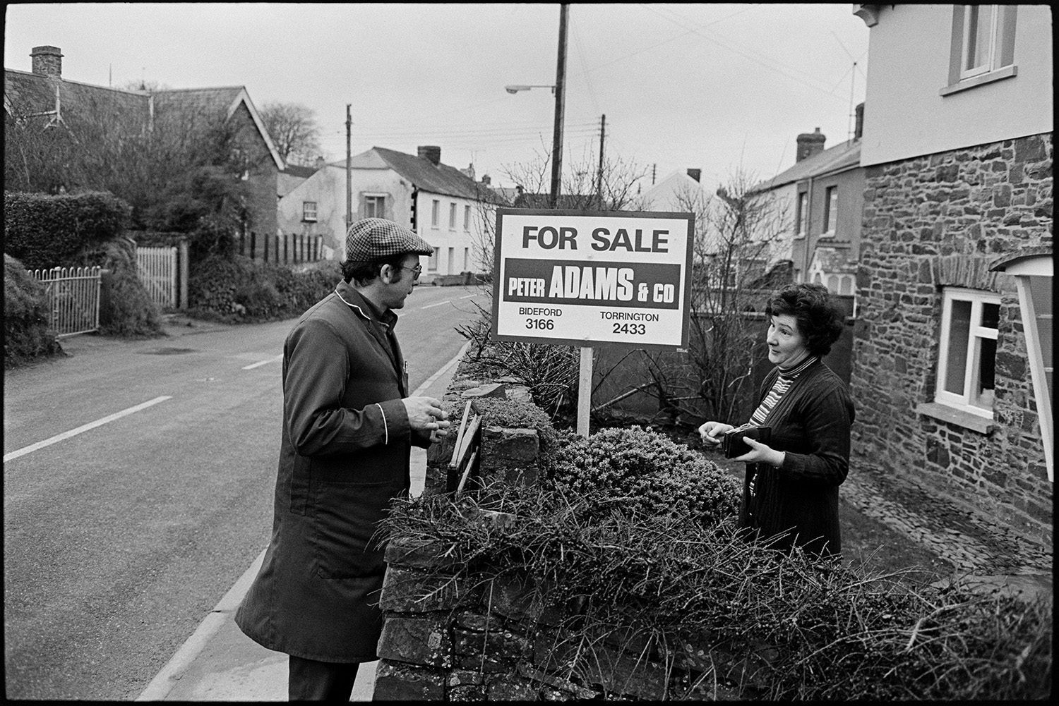 Farmer, milkman doing last milk round, showing new man the ropes, milk bottles.
[A woman standing in the front garden of her house in Beaford paying a milkman who is standing on the pavement at the front gate. A Peter Adams & Co for sale sign is in the front garden. Other houses can be seen in the street in the background.]