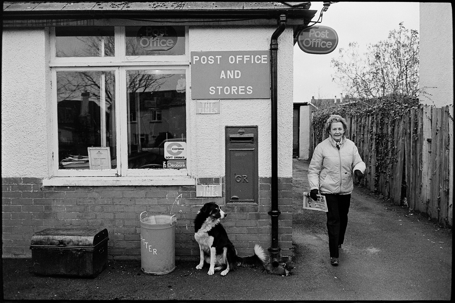 Milkman farmer on his last round in village, chatting as he delivers bottles from van.
[Woman holding a newspaper leaving Beaford Post Office and Stores. A post box is mounted in the wall and a dog, litter bin and a metal trunk are outside the shop window.]