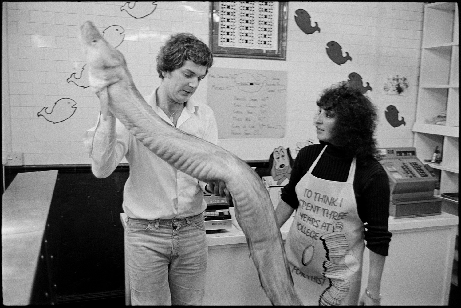 Fishmongers with giant conger eel ?? in shop. 
[A man in a fishmonger's shop in Allhaland Street, Bideford showing a giant conger eel to a woman wearing an apron with the wording 'To think I spent three years at college for this!']
