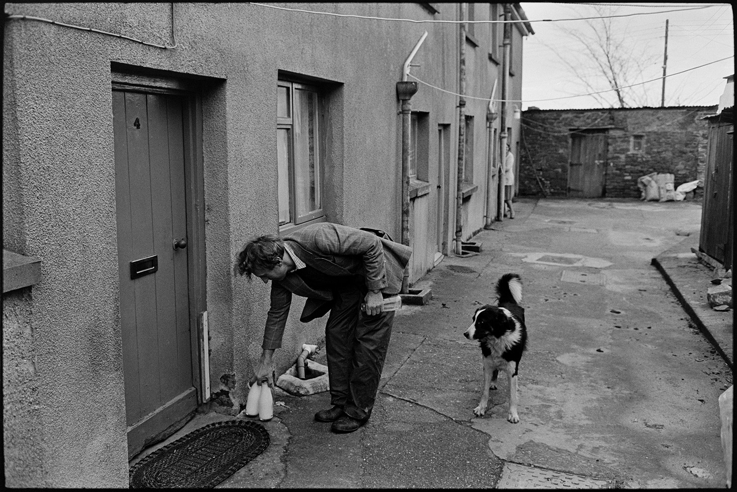 Milkman farmer on his last round in village, chatting as he delivers bottles from van.
[Ivor Bourne of Jeffrys Farm placing two bottles of milk in front of a house in Beaford. A dog is watching him and terrace of houses is visible in the background.]