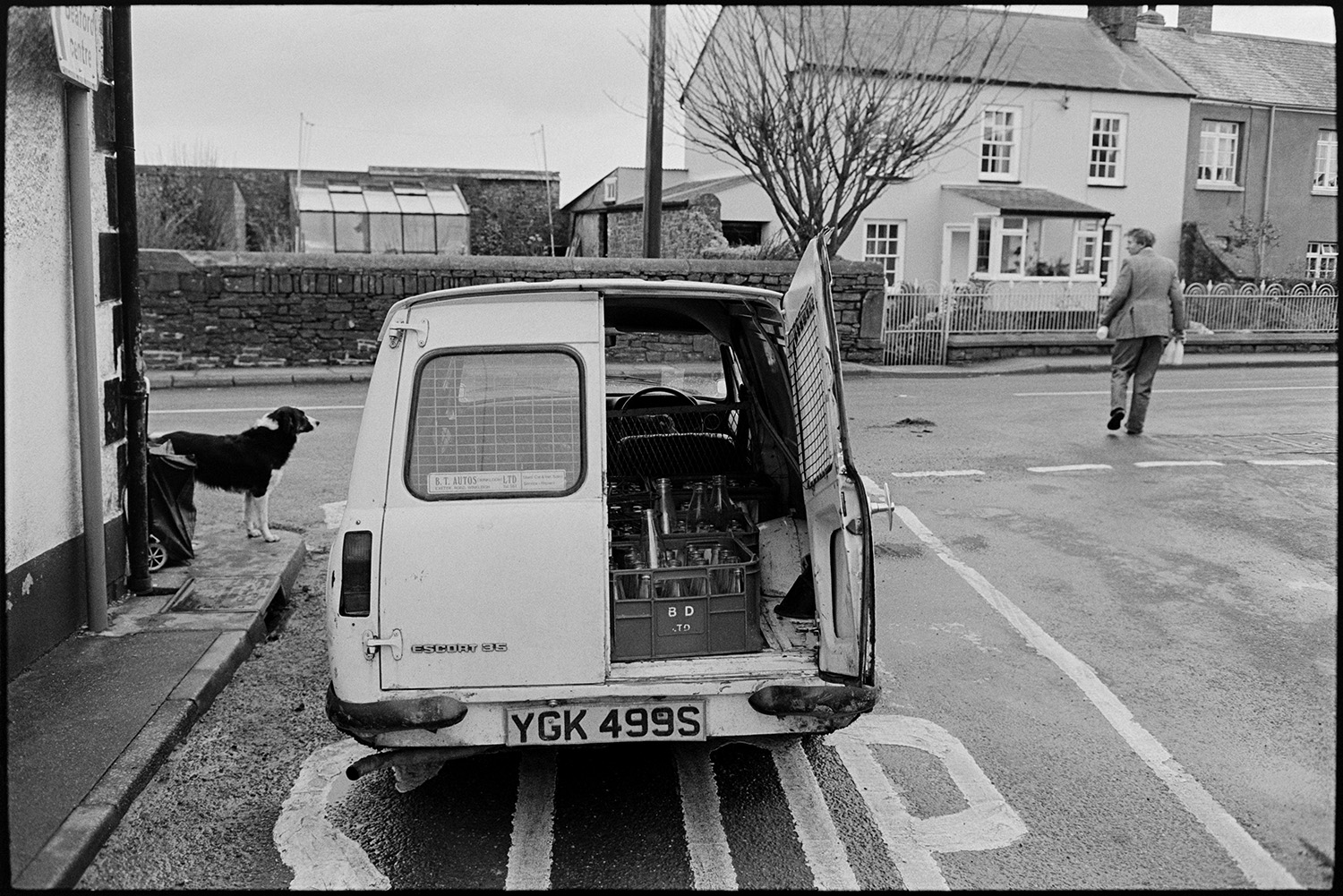 Milkman farmer on his last round in village, chatting as he delivers bottles from van.
[Ivor Bourne of Jeffrys Farm crossing a road in Beaford to deliver milk from his van on his last milk round. He is being watched by a dog standing on the corner of the street. Crates of empty milk bottles can be seen in the back of his van in the foreground.]