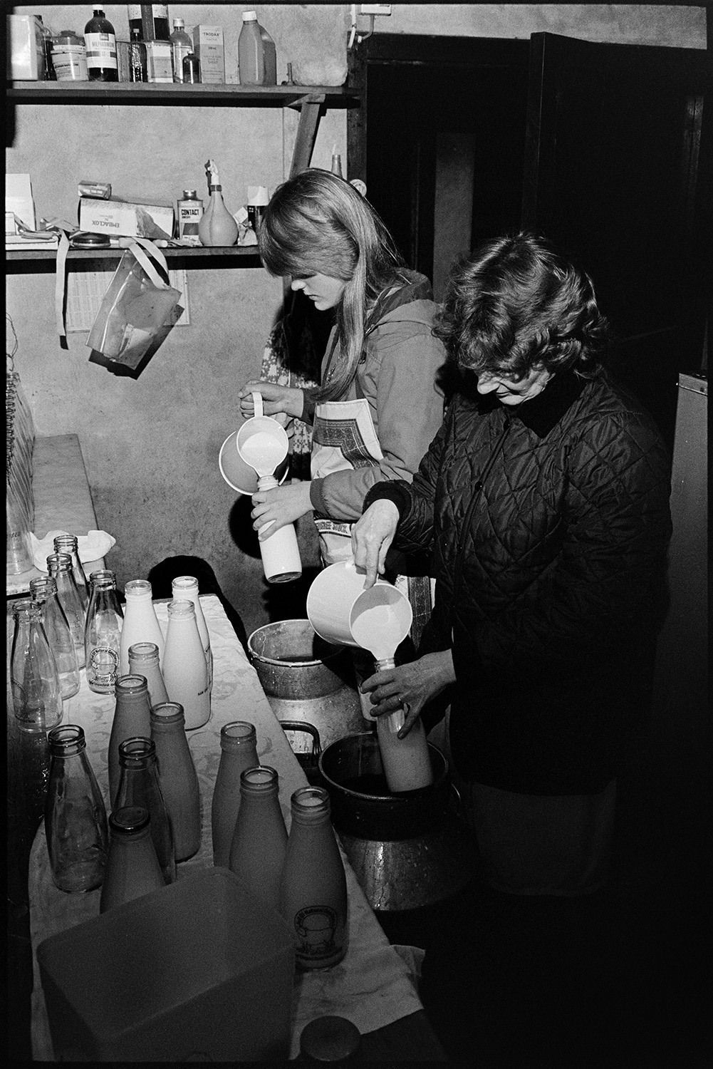 Women in dairy filling milk bottles, early morning.
[Two women, one of which is possibly Marylyn Bourne, filling milk bottles with milk from jugs in the dairy at Jeffrys Farm, Beaford, also known as Mill Road Farm.]