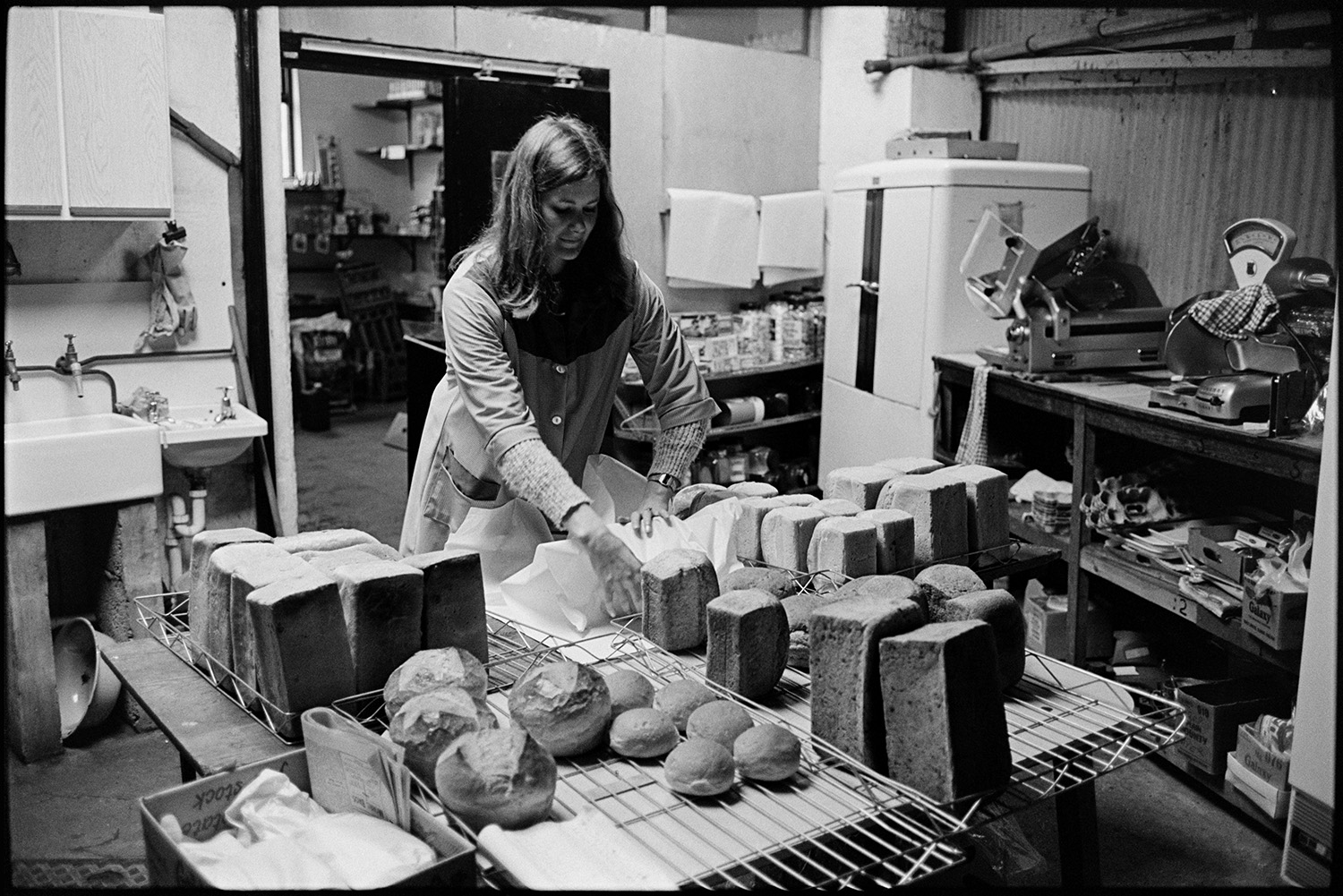 Women shop assistants wrapping bread in village shop store.
[Margaret Stoneman wrapping bread in the Church Street Stores, Dolton. Loaves and rolls are on the table in front of her and in the background a sink and scales are visible.]