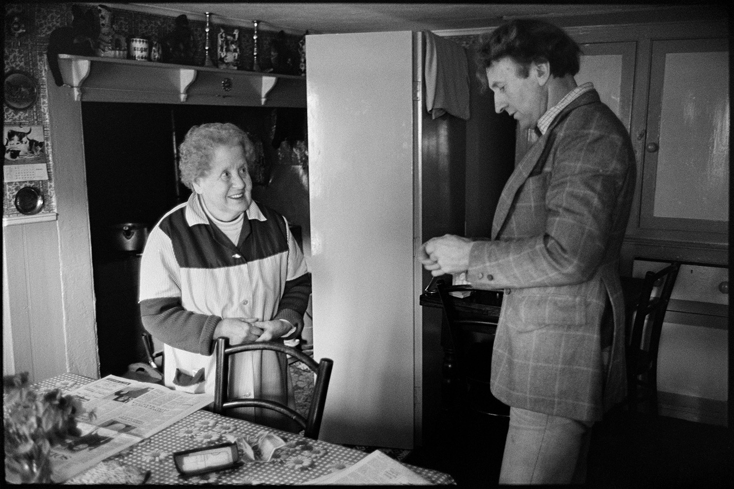 Milkman on last round chatting to customers, settling bills. Front of Post Office. 
[Ivor Bourne talking to a woman in her kitchen in Beaford after delivering her milk on his last milk round. Vases and candlestick are displayed on the mantelpiece in the background.]