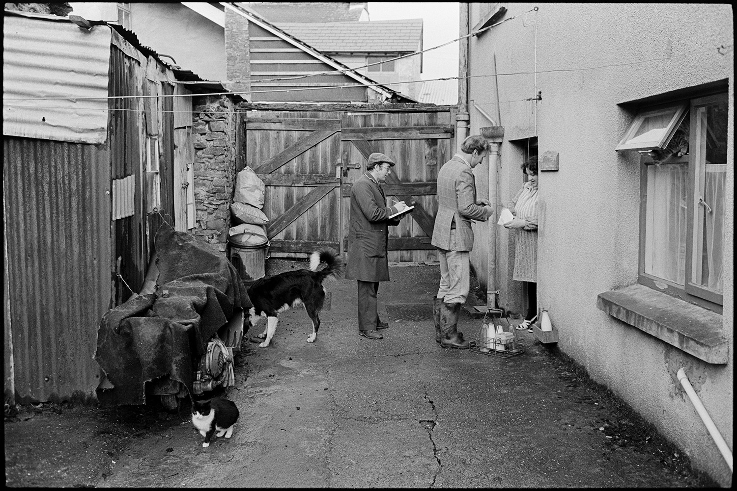 Milkman on last round chatting to customers, settling bills. Front of Post Office. 
[Ivor Bourne delivering milk to a woman at her house in Beaford on his last milk round. She is paying him for the milk. A man behind him is taking notes to take over the milk round. Also in the yard are a dog and a cat, and a corrugated  iron shed, opposite the house.]