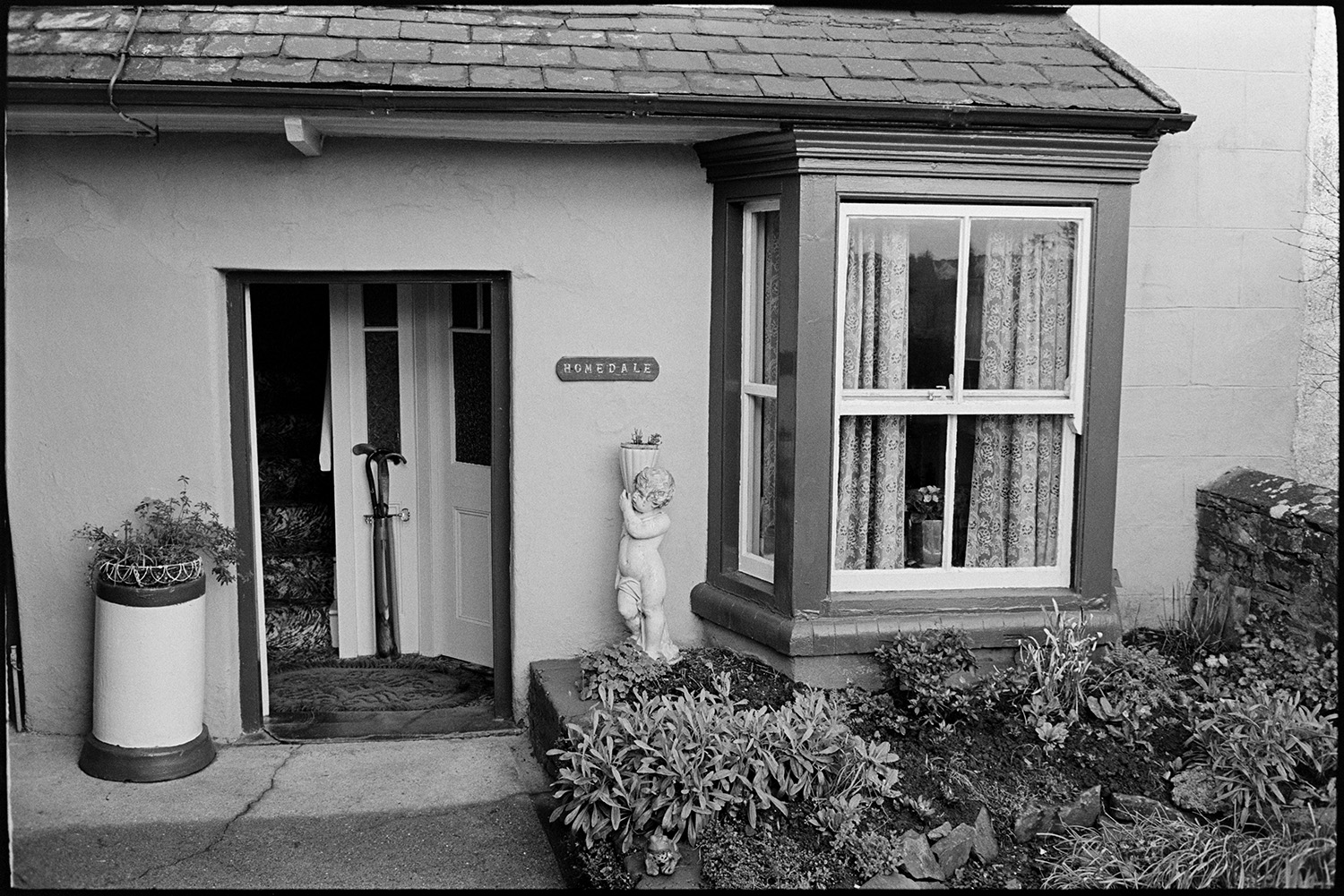 Milkman on last round chatting to customers, settling bills. Front of Post Office. 
[The front entrance to a house in Beaford called 'Homedale'. A plant containers and statue are by the front door and walking sticks can be seen just inside the entrance.]
