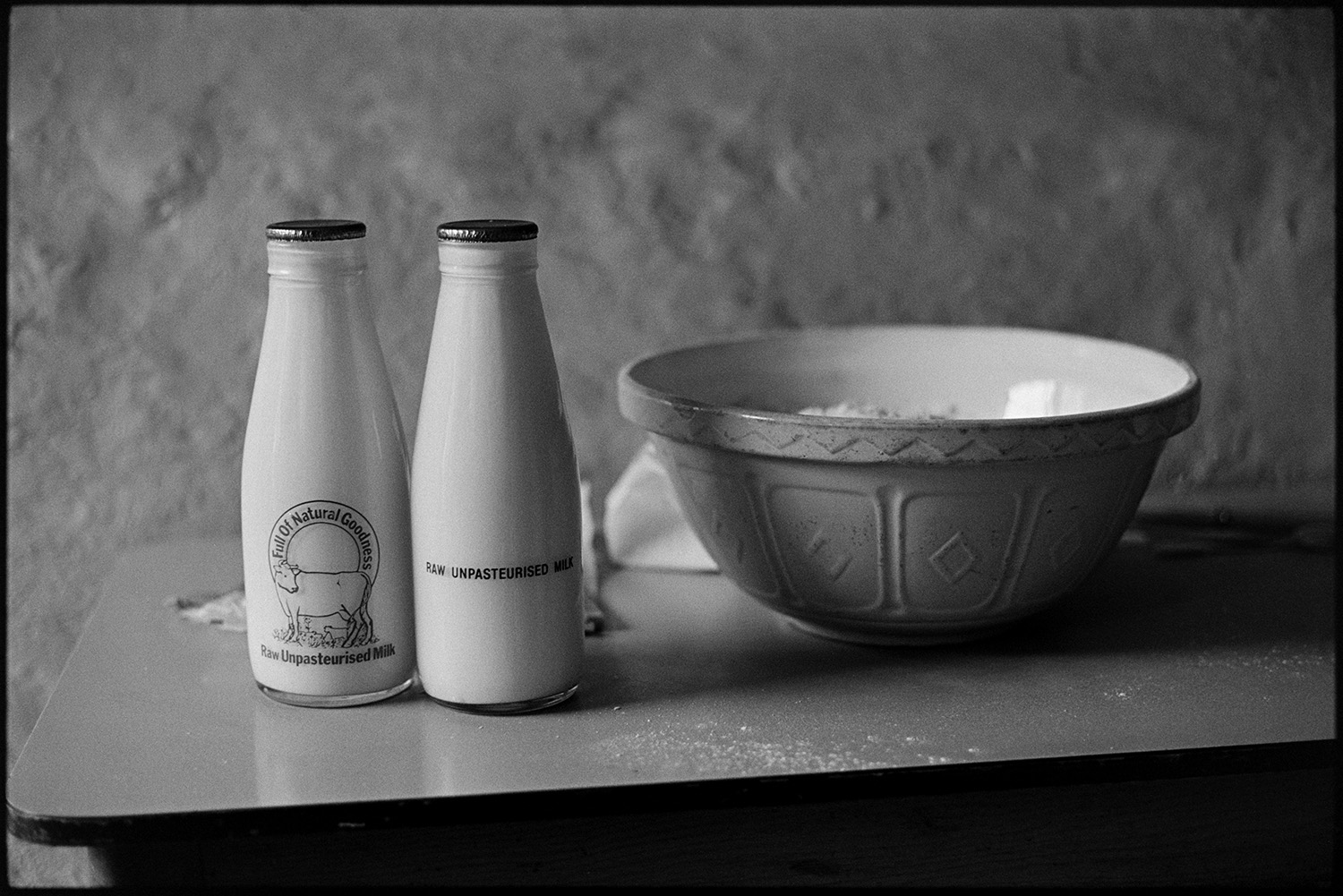 Milk bottles and pastry bowl on table at cottage door.
[Two full bottles of milk labelled 'Raw Unpasteurised Milk - Full of Natural Goodness' next to a mixing bowl  on a table at Chaplands, Cottage, Beaford.]