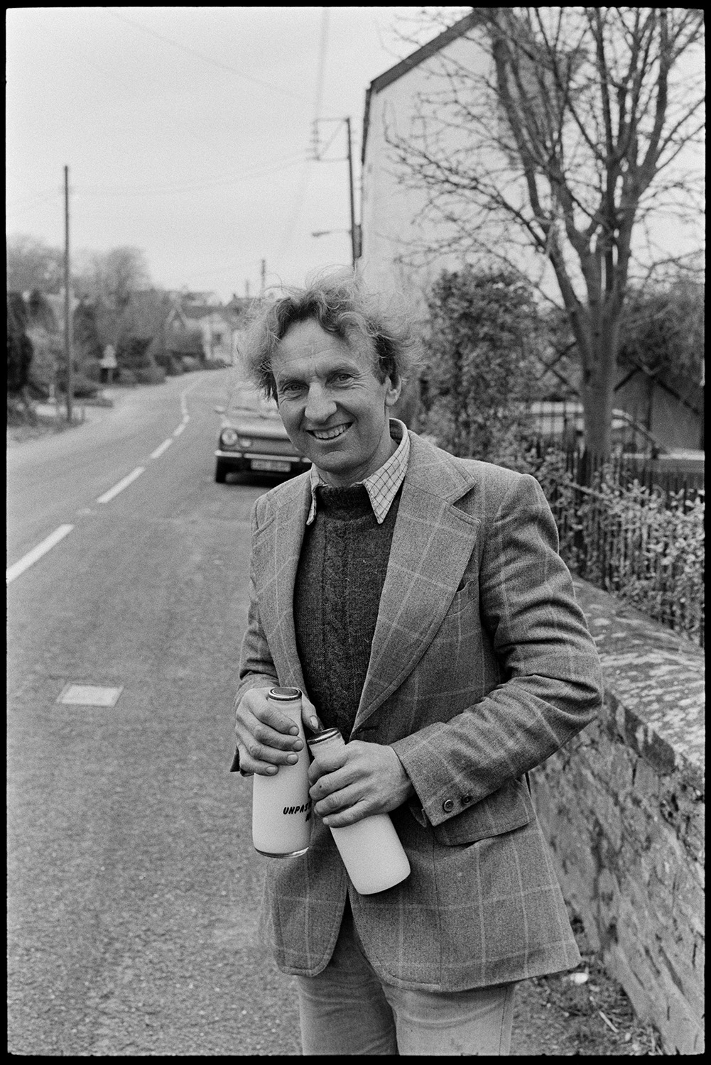 Milkman on last round.
Ivor Bourne standing in road in Beaford holding two full bottles of milk on his last milk delivery round in the village.]