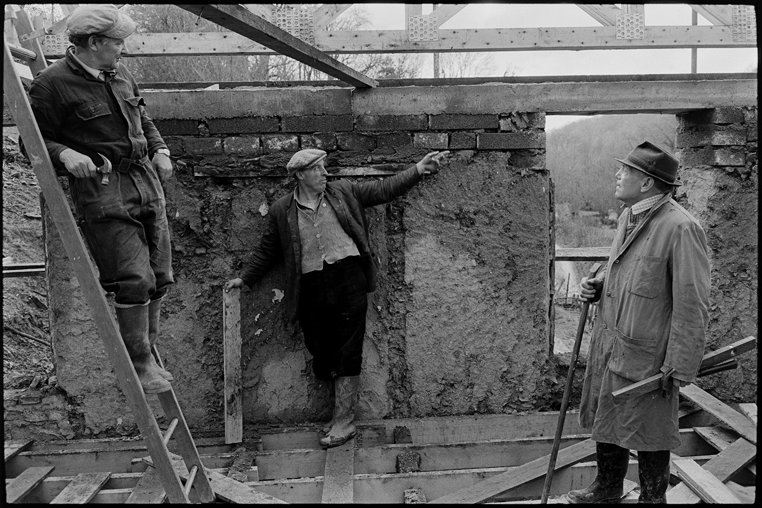 Builder renovating cottage with new roof.
[Builders renovating a cottage at Millhams, Dolton. Cob walls, concrete lintels, wooden rafters and joists are shown. One man is pointing at the roof, another builder is standing on a ladder and holding a hammer, while the third man is carrying a spirit level and hammer.]