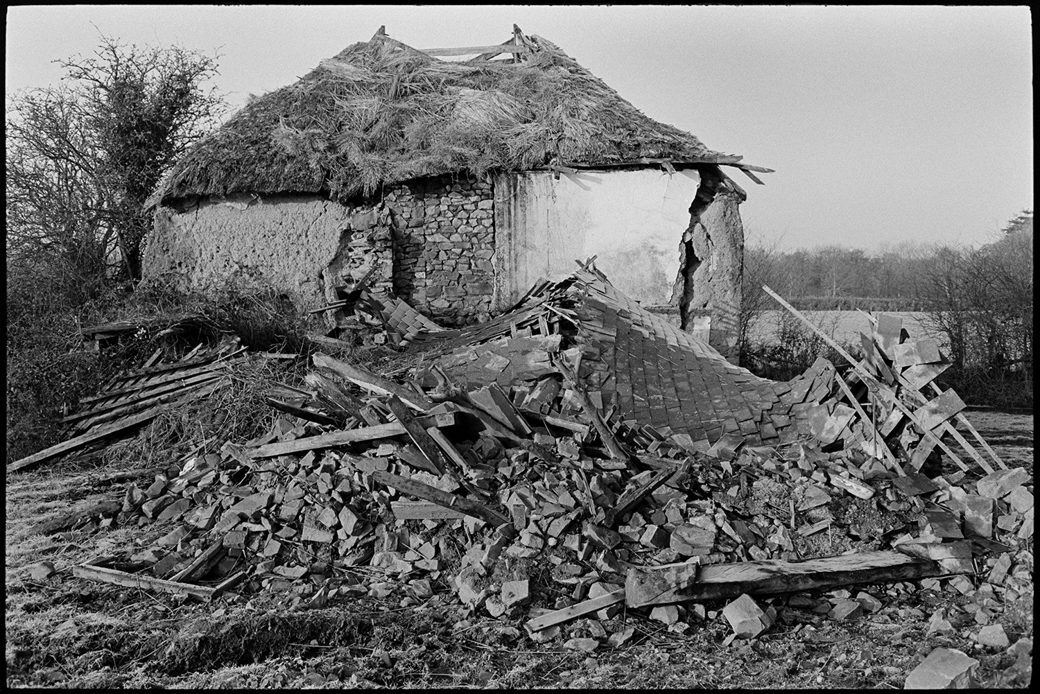 Ruined thatch and cob barns, with views from nearby stream, trees.
[A ruined cob and thatch barn in a field at Middle Week, Iddesleigh, behind a pile of rubble including slates and timber.]