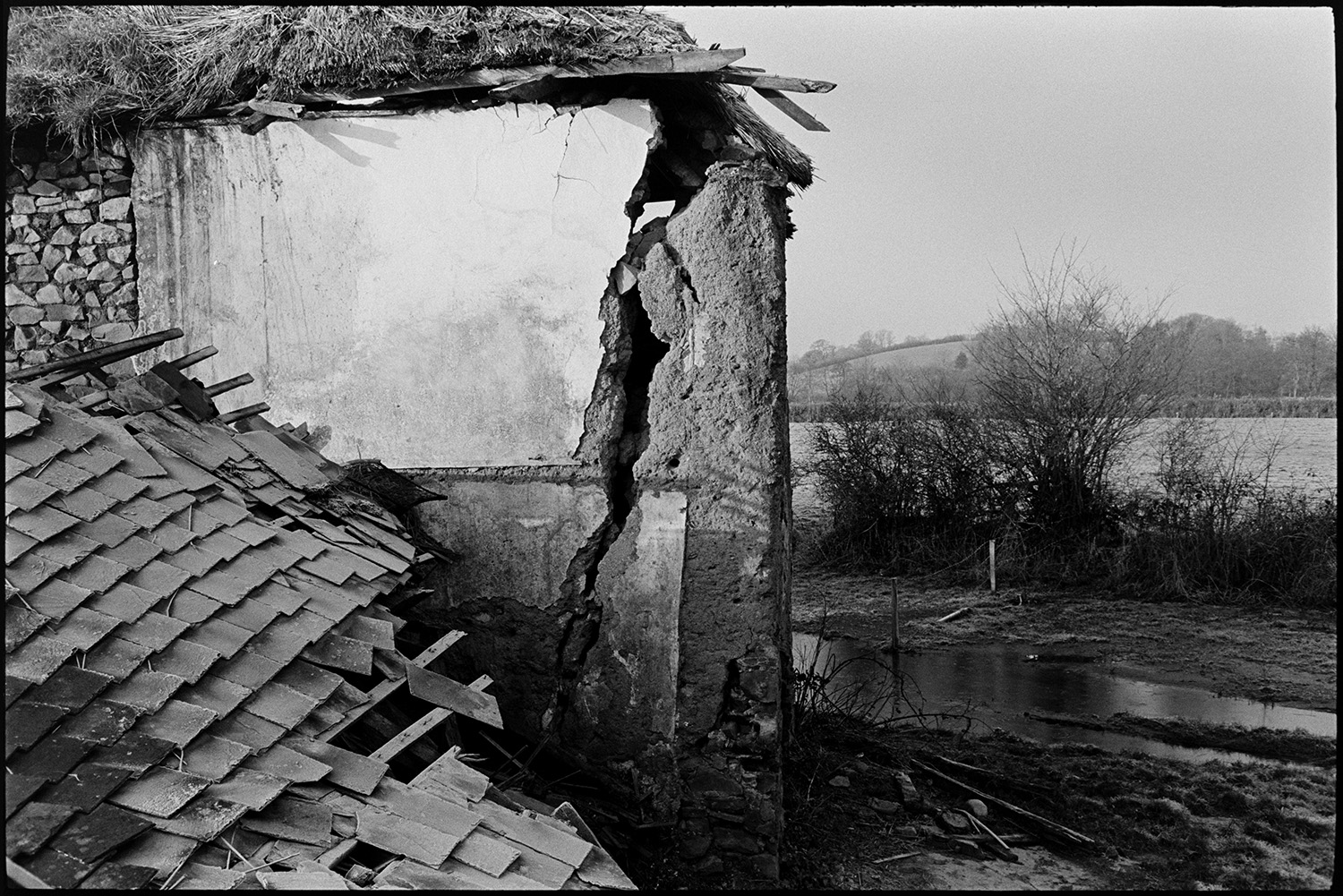 Ruined thatch and cob barns, with views from nearby stream, trees.
[A ruined cob and thatch barn at Middle Week, Iddesleigh, behind a pile of fallen slates and timbers. A large crack can be seen in the barn wall. A waterlogged field and hedges are visible in the background.]