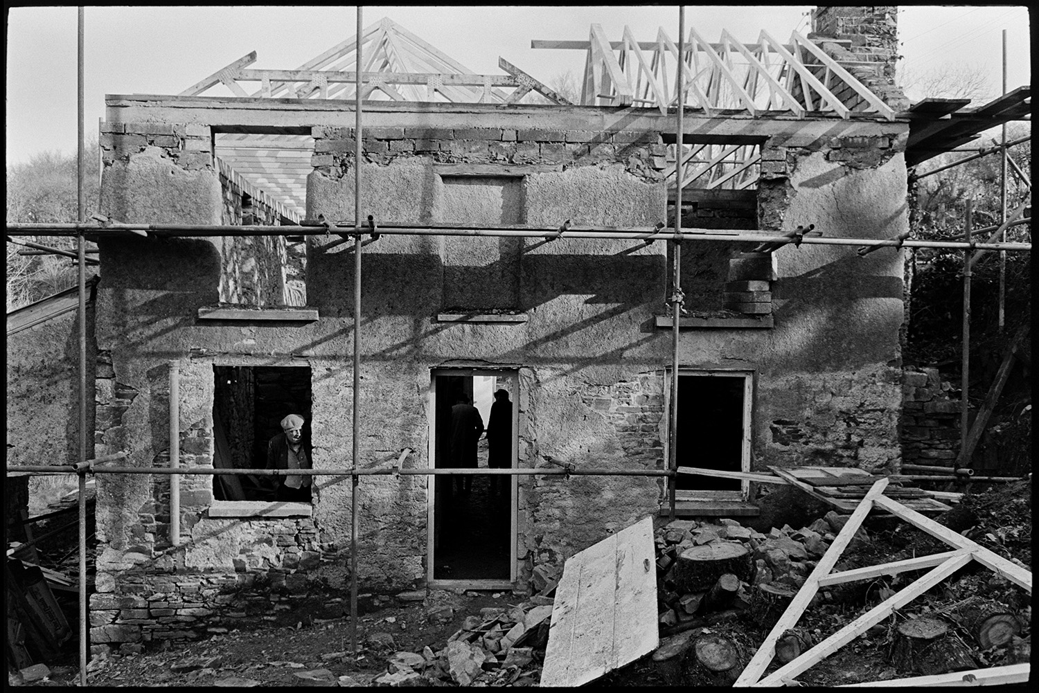 Man rebuilding roof of house another looking on, scaffolding. View of front of house.
[George Ayre looking out of a cottage window at Millhams, Dolton during renovation work. Wood roof timbers and cob walls are visible behind scaffolding, with building debris on the ground.]