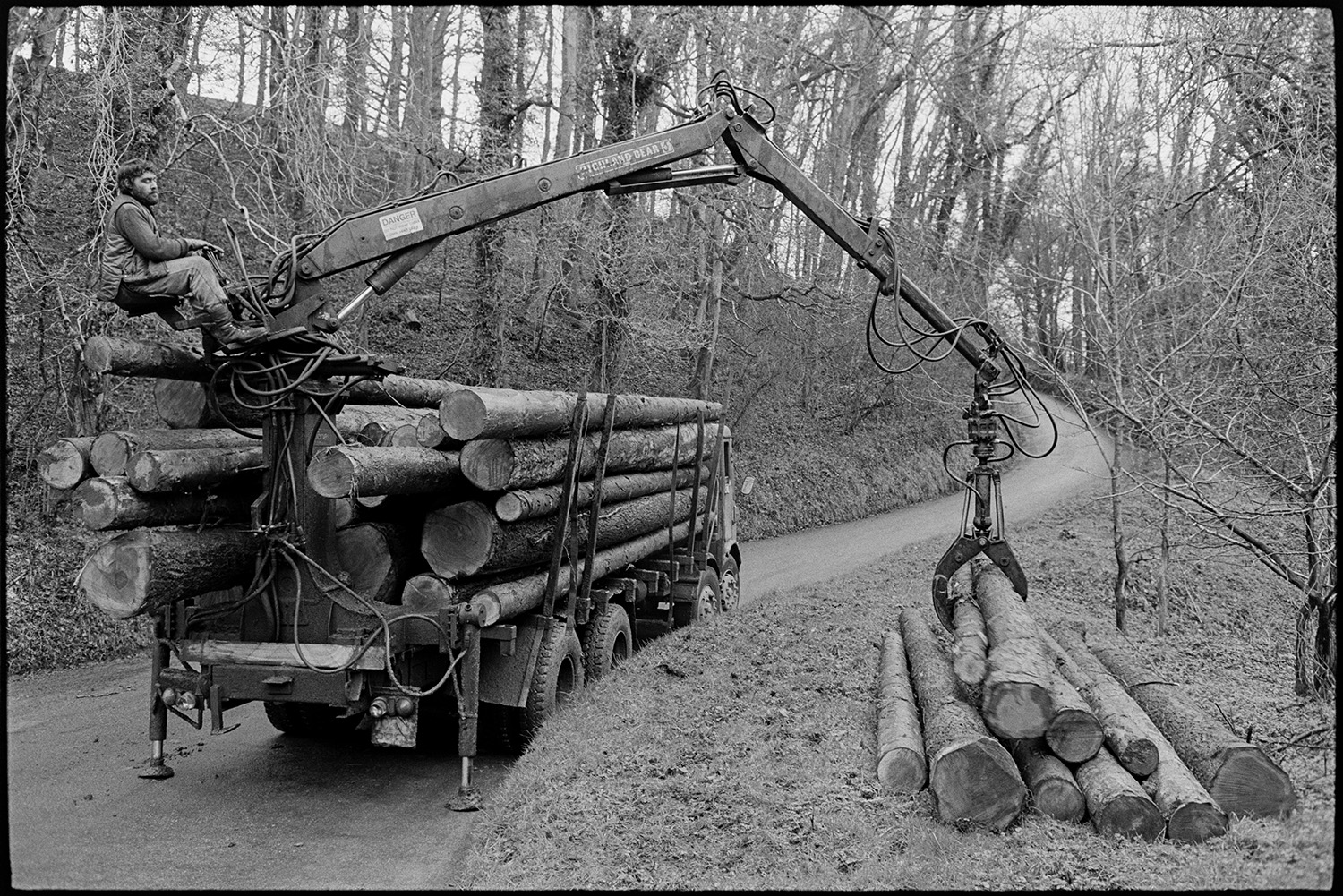 Man loading logs onto timber lorry. 
[A man operating a winch to load logs or timber onto a lorry in woodland at Cleave, Dolton.]