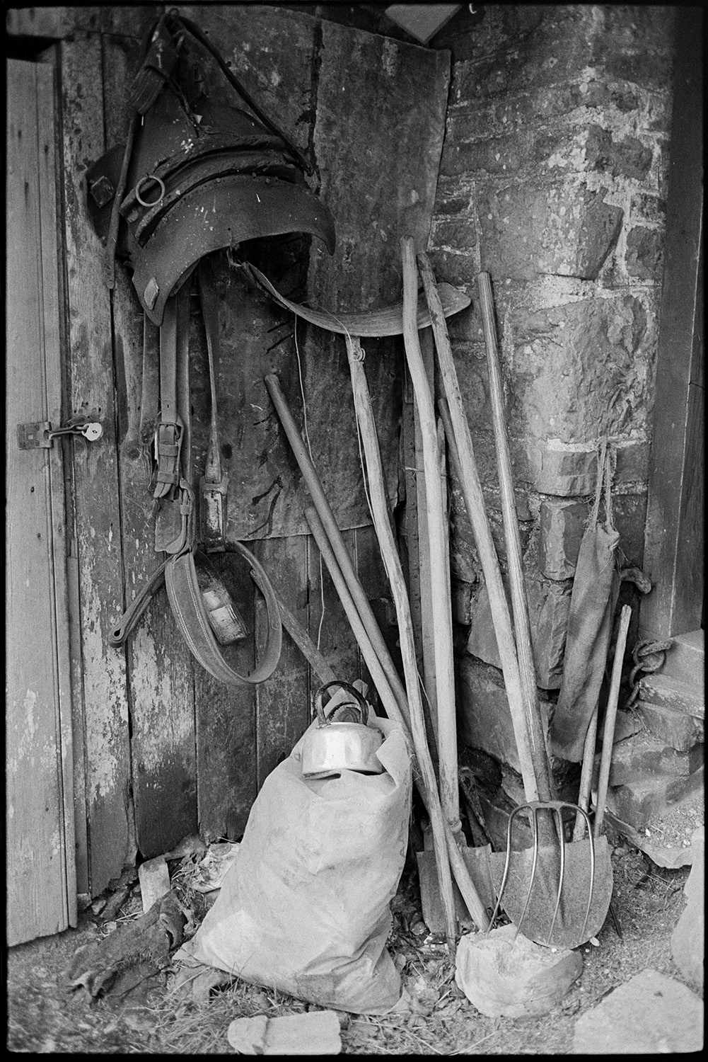 Implements in corner of farm shed, three pronged fork. 
[Spades, a fork, a sack, leather straps and possibly a saddle stacked in the corner of a farm shed by a wooden door, at Colehouse, Riddlecombe.]