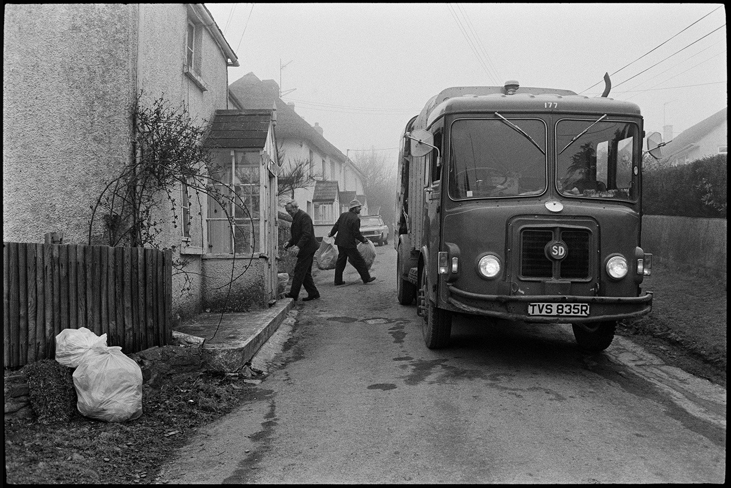 Refuse dust cart, lorry, village street, men loading. Dustmen. 
[Binmen collecting rubbish bags from outside houses in West Lane, Dolton and loading them into a bin lorry.]