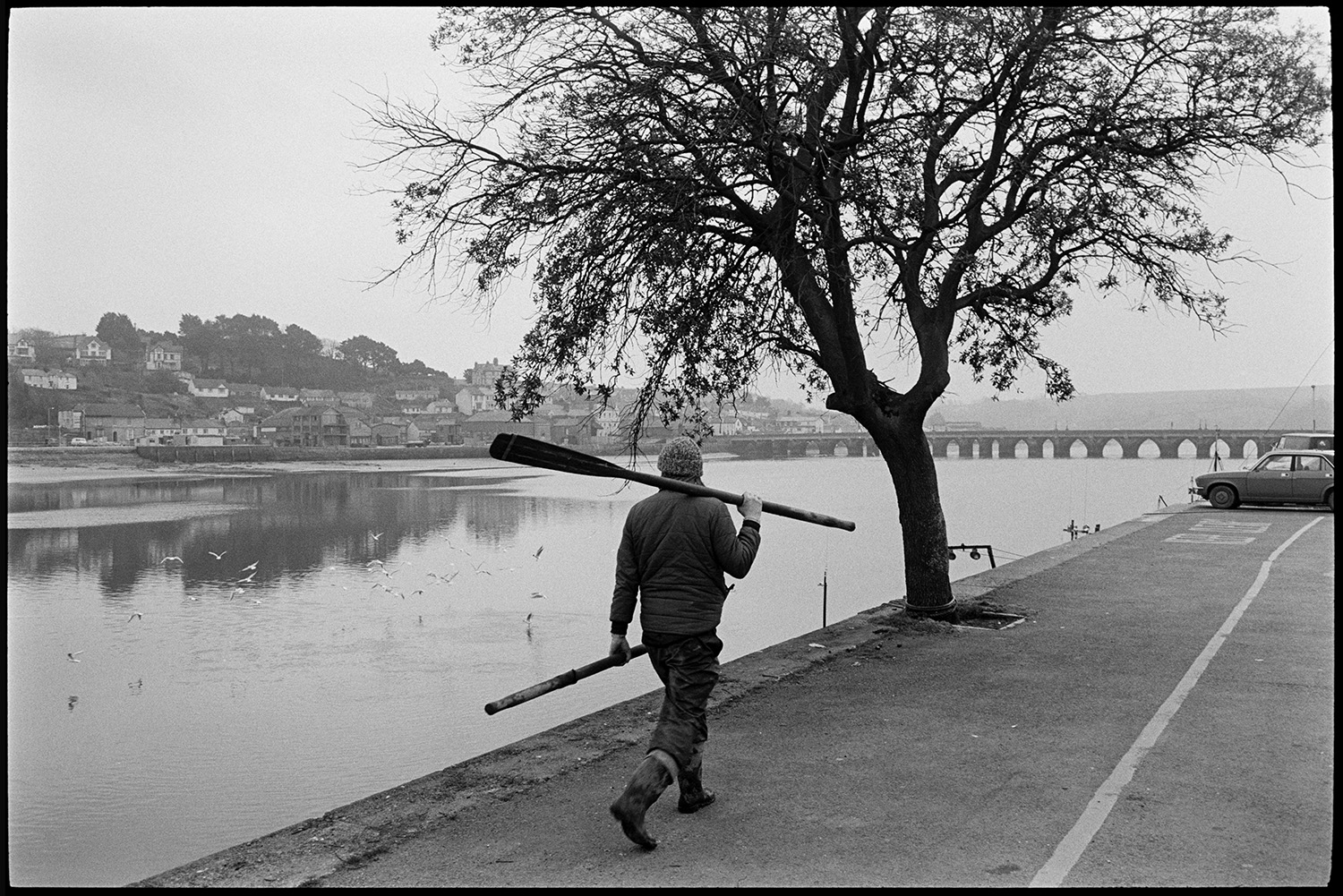 River and quay, oarsmen, fishermen mending nets, old bridge in background, fishing boats. 
[A person walking along Bideford Quay carrying two oars. They are passing a tree. Bideford old bridge can be seen in the background and houses are visible on the opposite side of the River Torridge.]