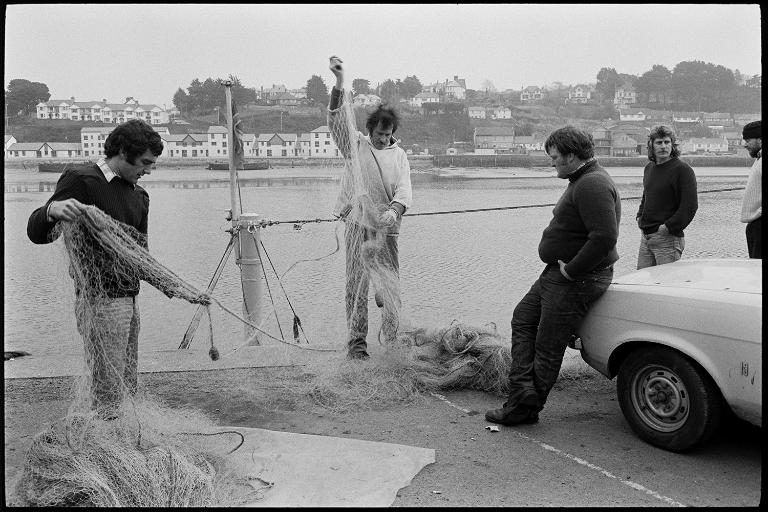 River and quay, oarsmen, fishermen mending nets, old bridge in background, fishing boats. 
[Fishermen sorting and mending their fishing nets on Bideford Quay. The River Torridge and houses on the opposite side of the river can be seen in the background.]