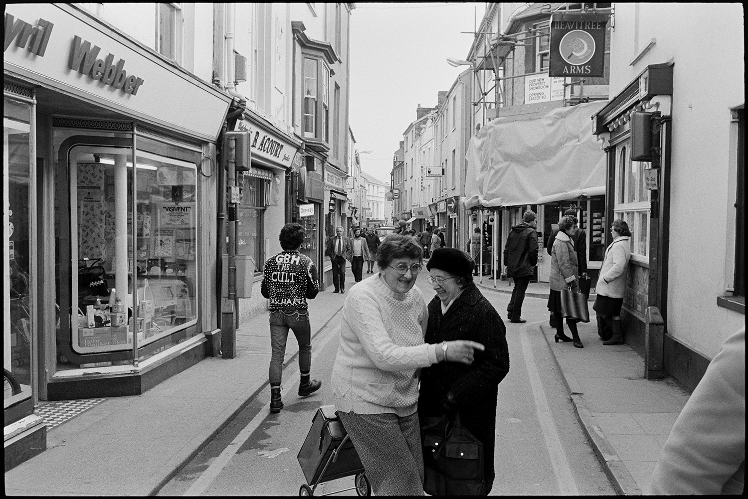 Street scenes, people chatting outside pub. 
[Two women talking and laughing in the street outside the Heavitree Arms pub in Mill Street, Bideford. One of them is pulling a shopping trolley. Other shoppers can be seen in the street, including a person wearing a jacket with the logo of the Torrington punk rock bank, The Cult Maniax. Shop fronts of Cyril Webber and B A Court are visible.]