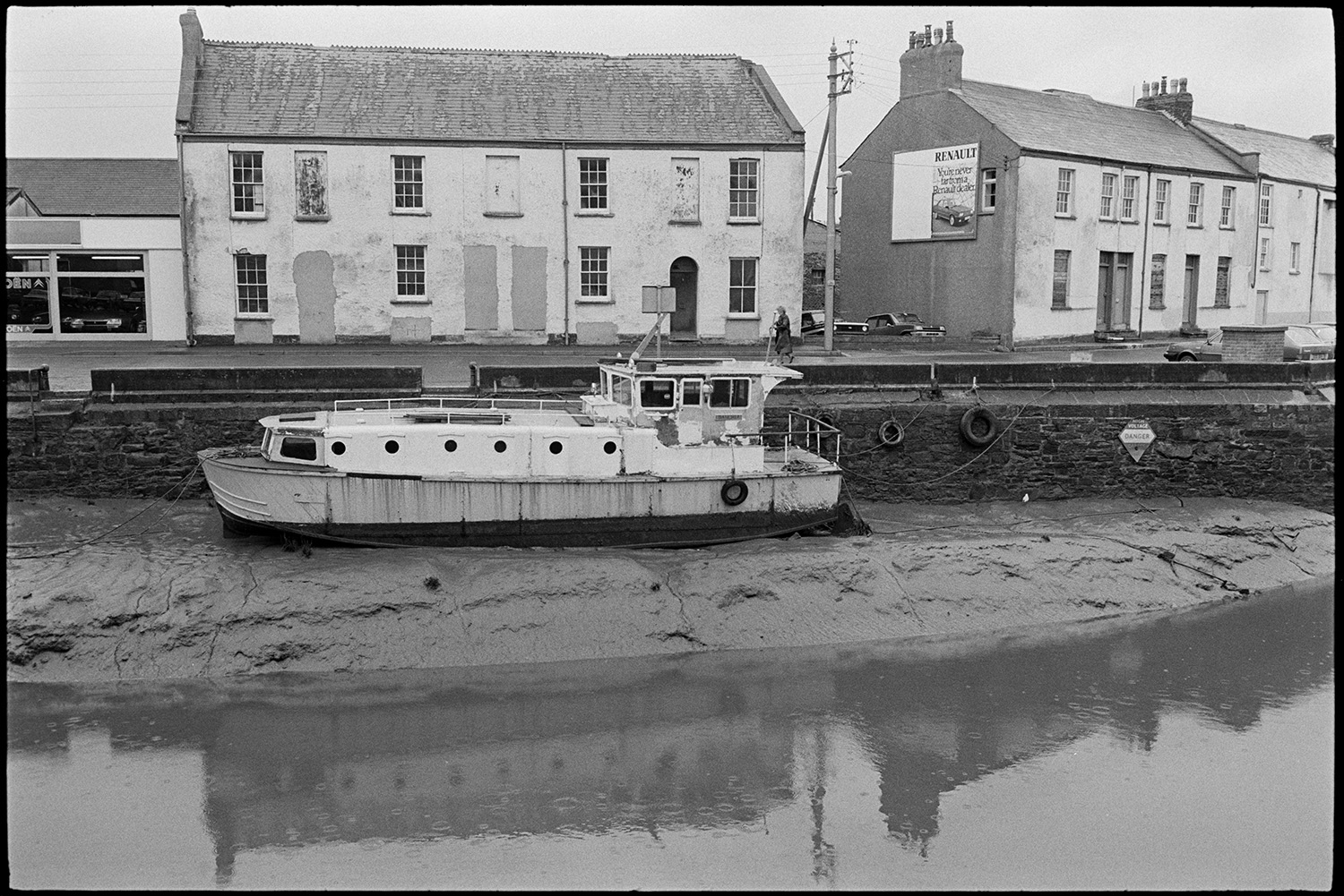Boat, barge at quayside, houses before renovation.
[View of a boat or barge moored on the mud bank by the Rolle Quay, Barnstaple. Houses behind the Quay with blocked up doorways are visible. They were later renovated.]